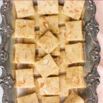Rectangular pieces of badam burfi are arranged on an ornate silver tray; text at the top reads “Quick & Easy Badam Burfi: 30 minute recipe.”.