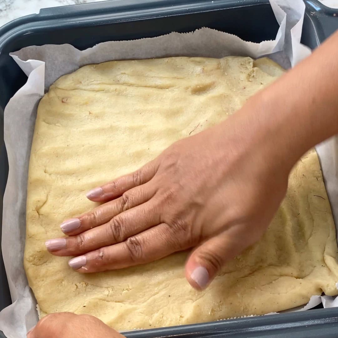 A hand presses dough evenly into a parchment-lined square baking pan.