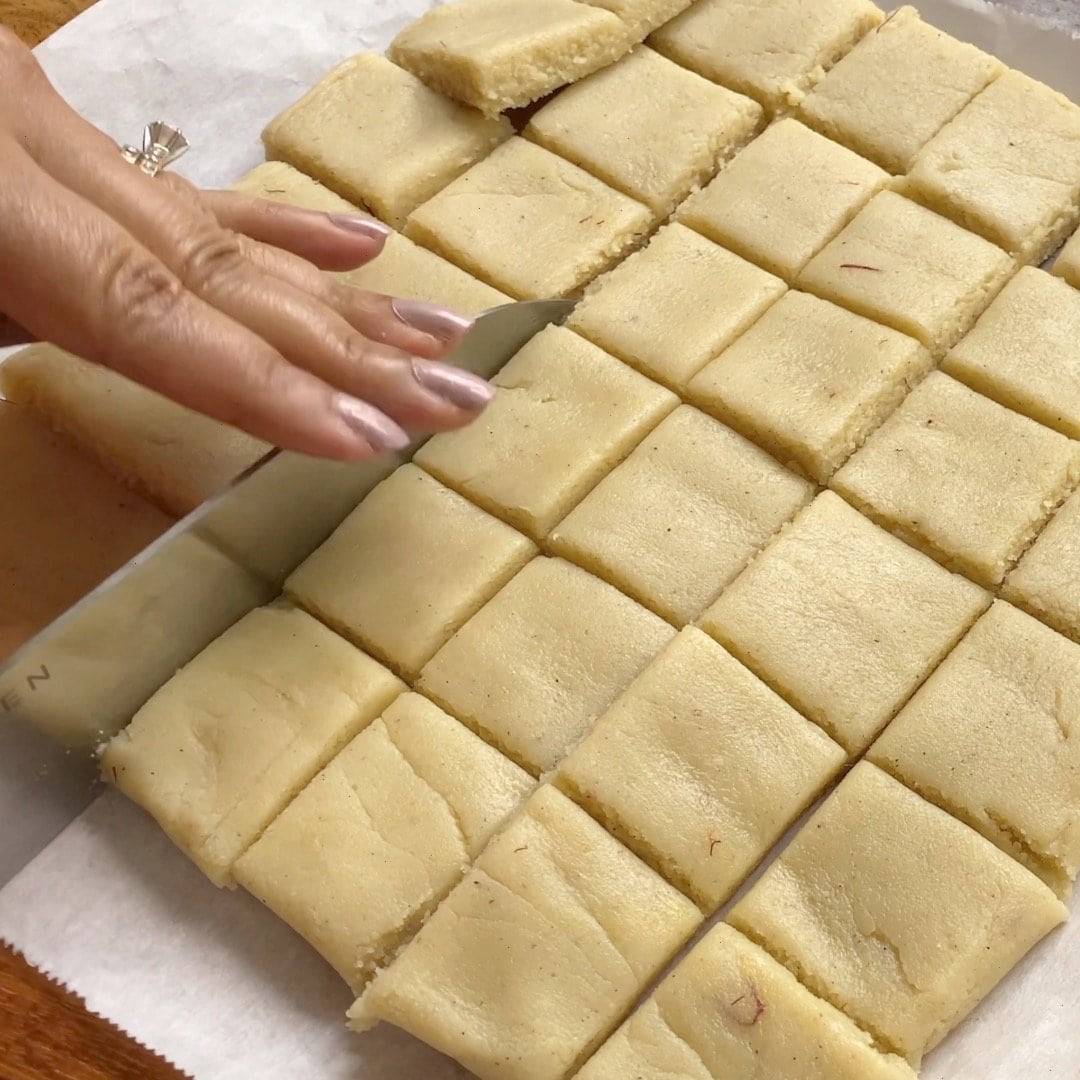 Cutting a tray of square-shaped burfi with a knife on parchment paper.