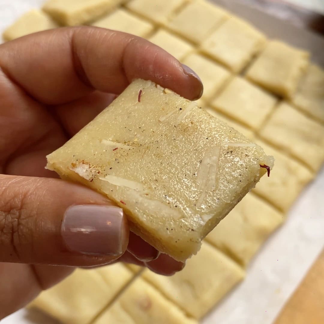 A hand holding a square piece of badam burfi.