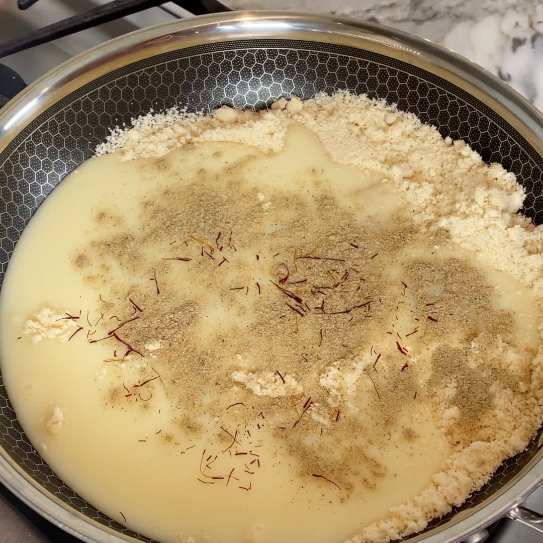 A frying pan contains flour, milk, saffron strands, and ground spices on a stovetop, before mixing and cooking.