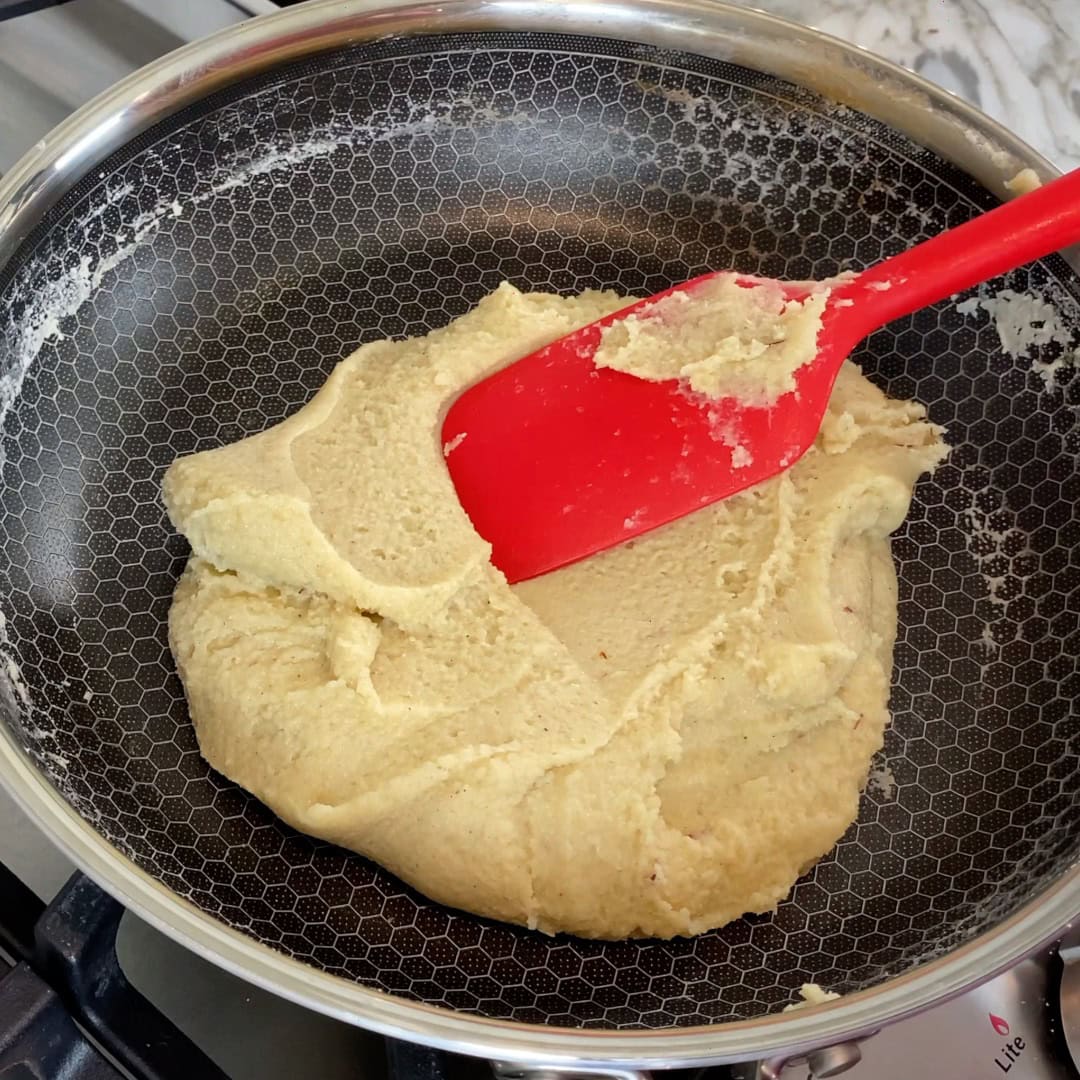 A red spatula stirs a thick, beige dough-like mixture in a frying pan on a stove.