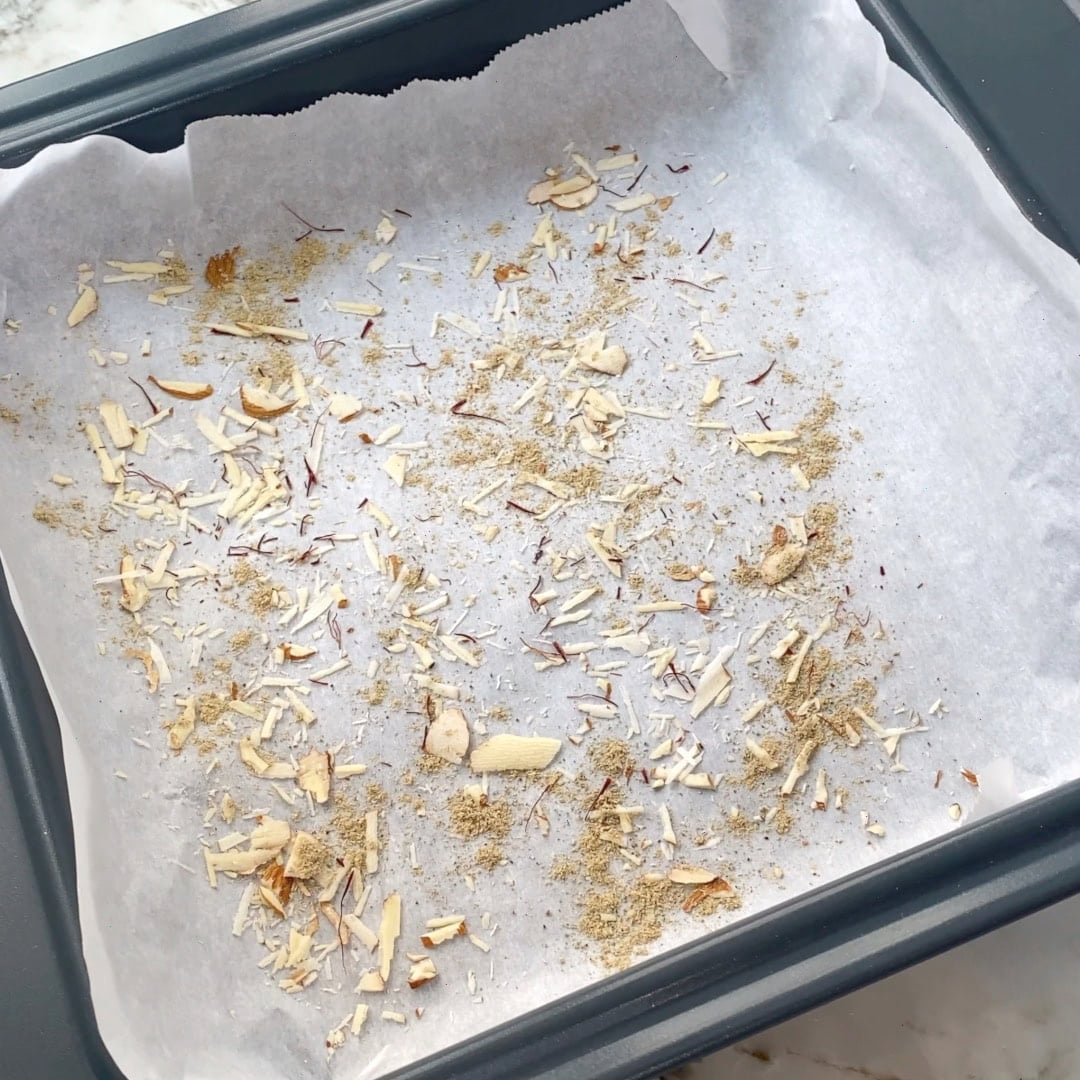 A baking pan lined with parchment paper, topped with scattered slivered nuts, saffron strands, and ground cardamom.