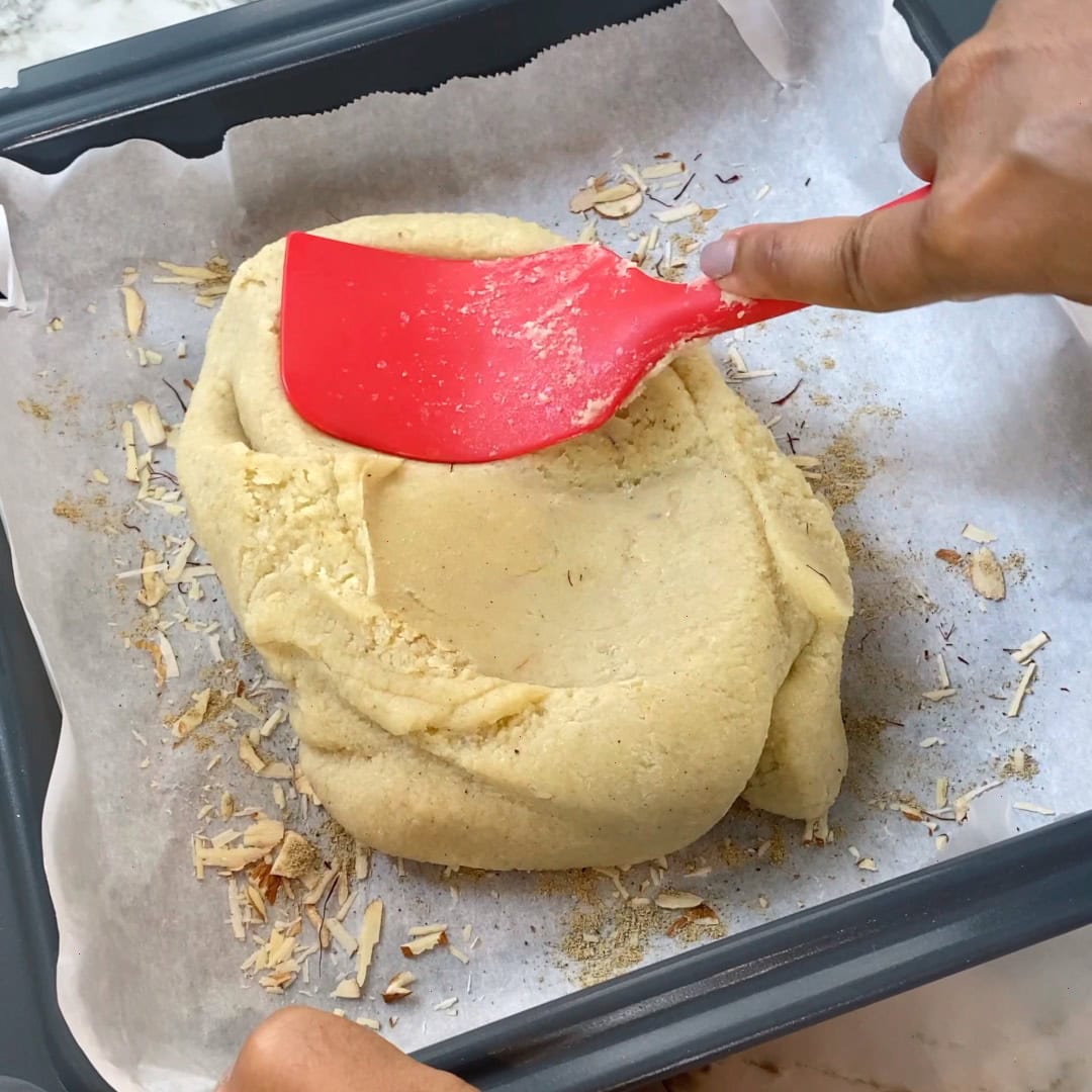 A person spreads dough in a parchment-lined baking tray using a red spatula; sliced almonds are scattered underneath the dough.
