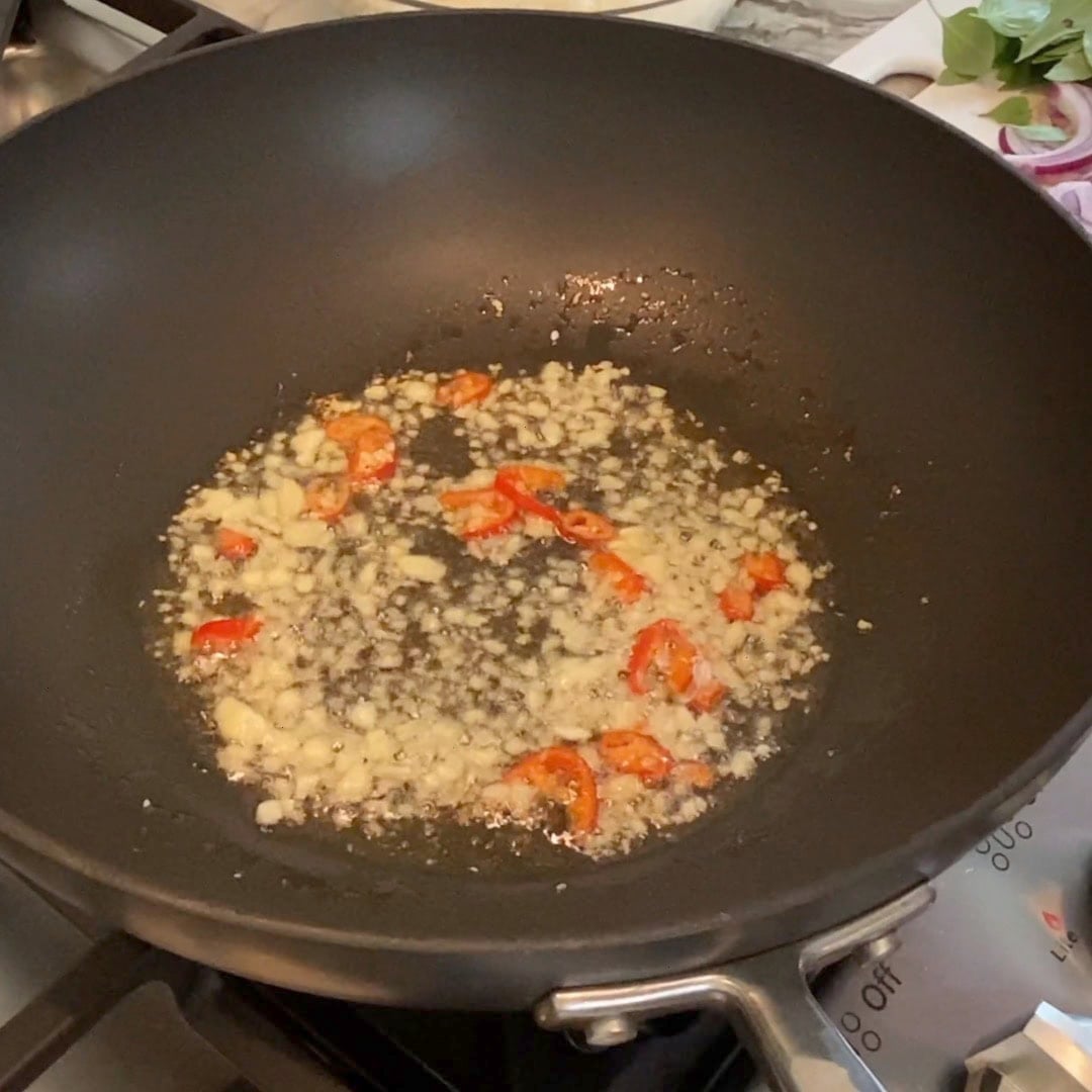 Chopped garlic and red chili slices sautéing in oil in a black wok on a stove.