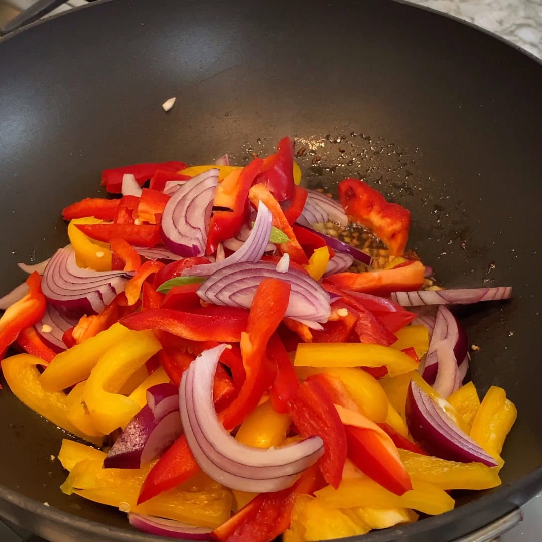 Sliced red onions, red bell peppers, and yellow bell peppers are cooking in a black wok.