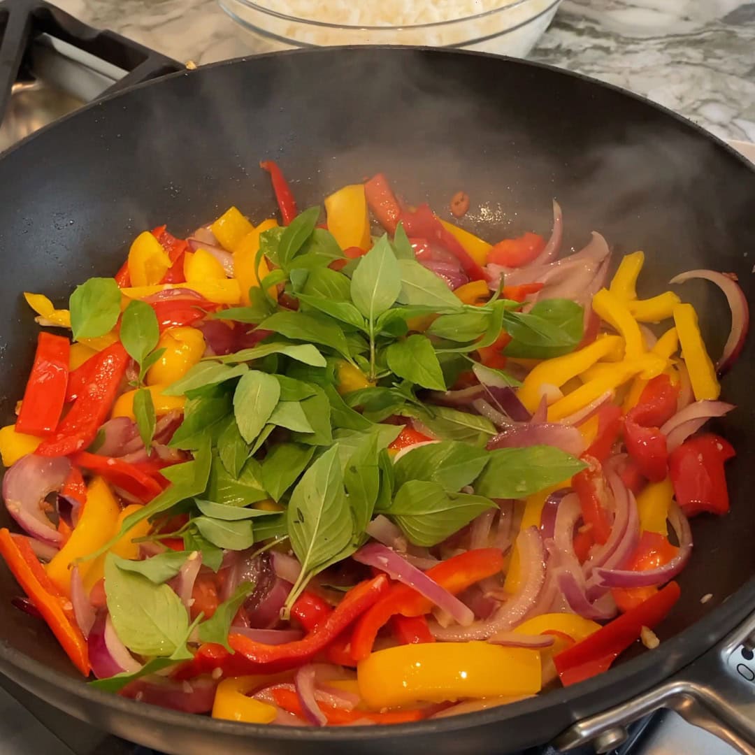 A wok with sliced red and yellow bell peppers, red onions, tomatoes, and fresh basil leaves being sautéed on a stovetop.