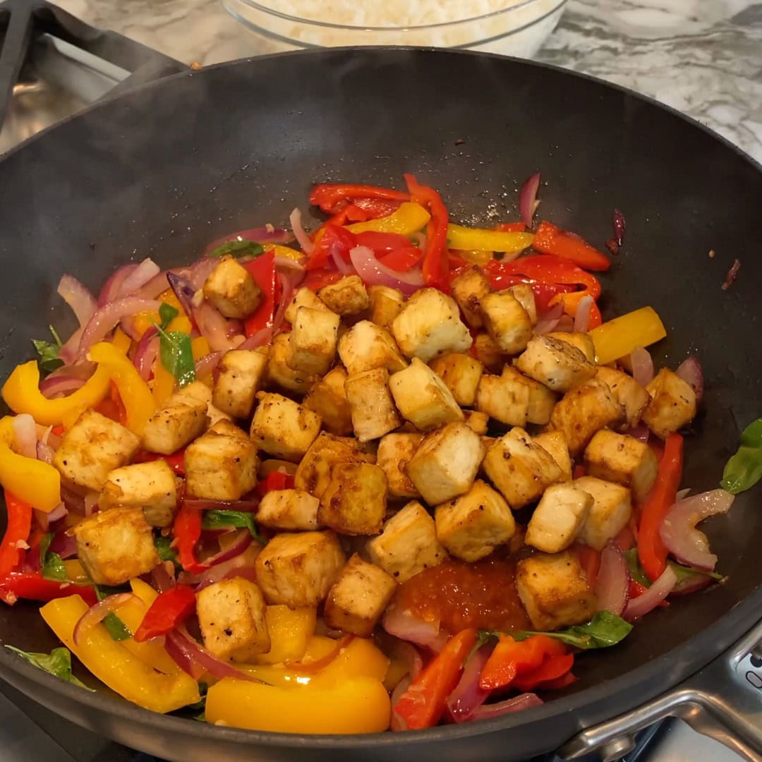 Cubed tofu and sliced red and yellow bell peppers, onions, and basil are being stir-fried in a pan on a stovetop.