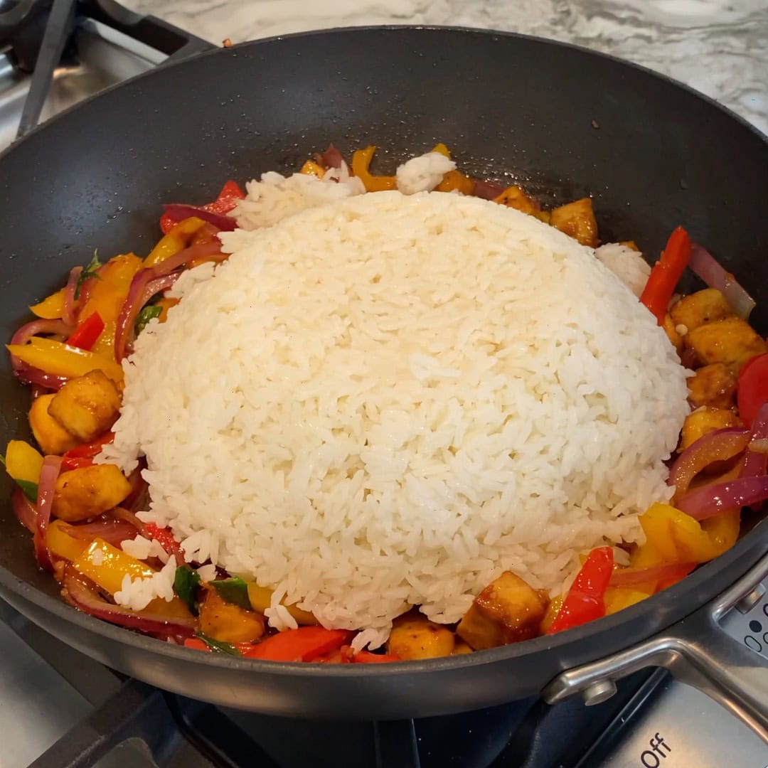 A mound of cooked white rice sits in the center of a pan surrounded by sautéed tofu, bell peppers, and red onions.