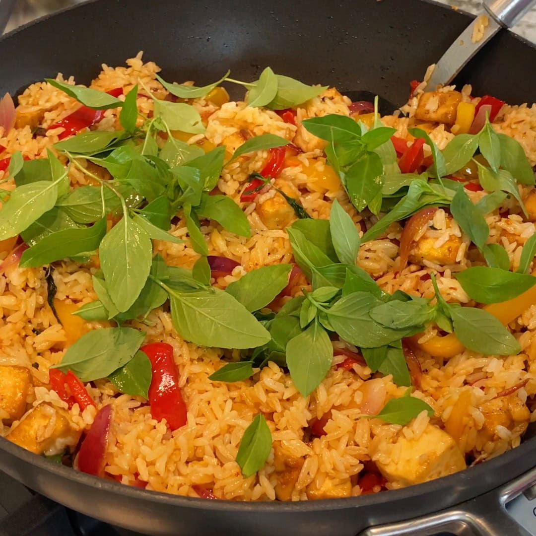 A pan of stir-fried rice with tofu, red and yellow peppers, onions, and topped with fresh basil leaves.
