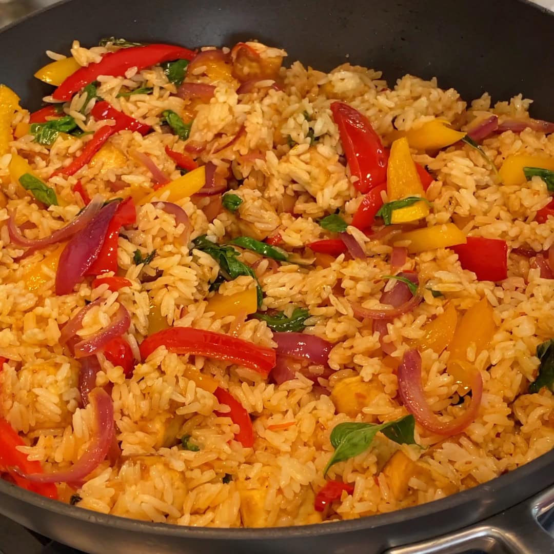 A skillet filled with stir-fried rice, red and yellow bell peppers, red onions, and fresh basil leaves.