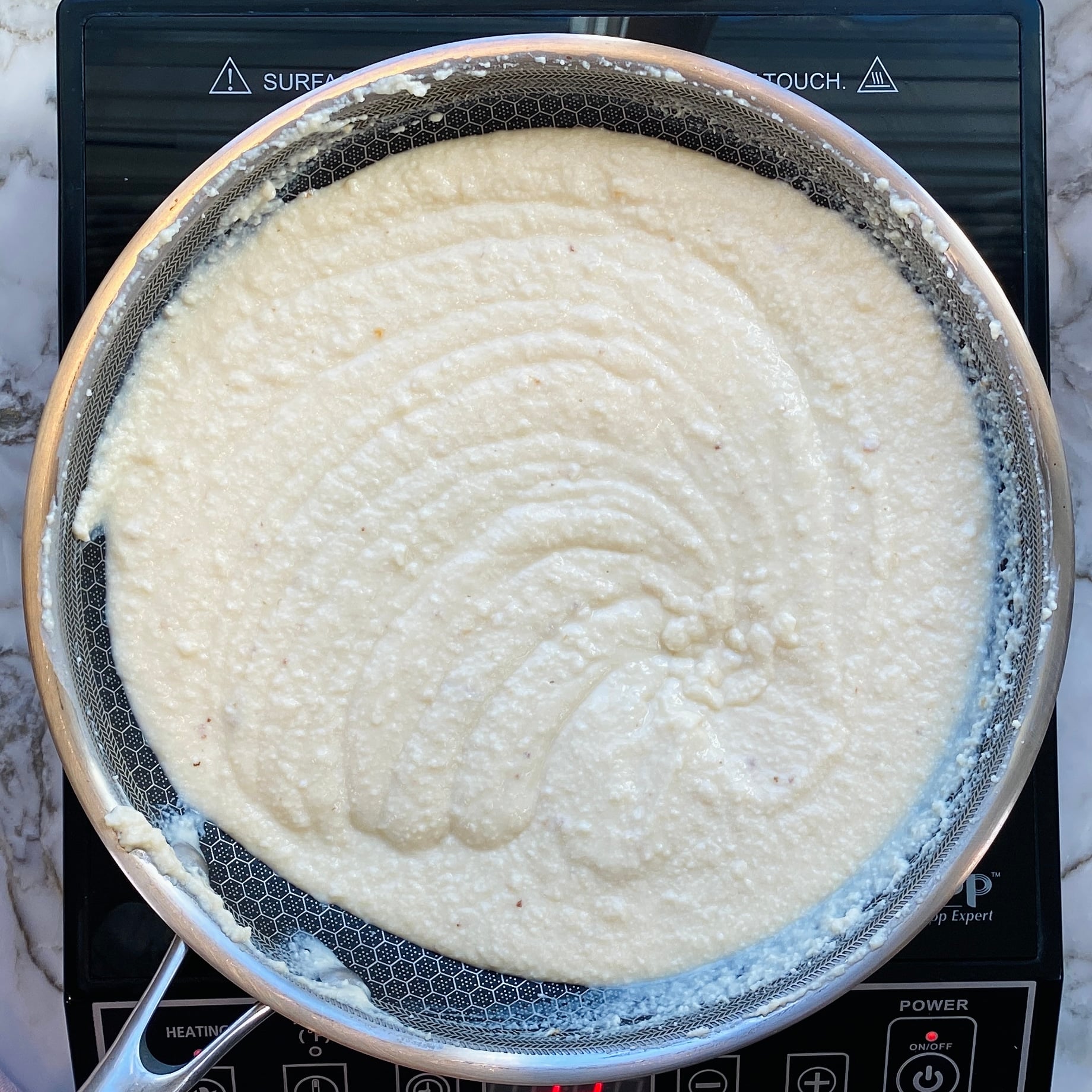 A pan filled with white, grainy batter is placed on an induction cooktop. The surface of the batter shows swirled patterns, indicating it has been stirred.