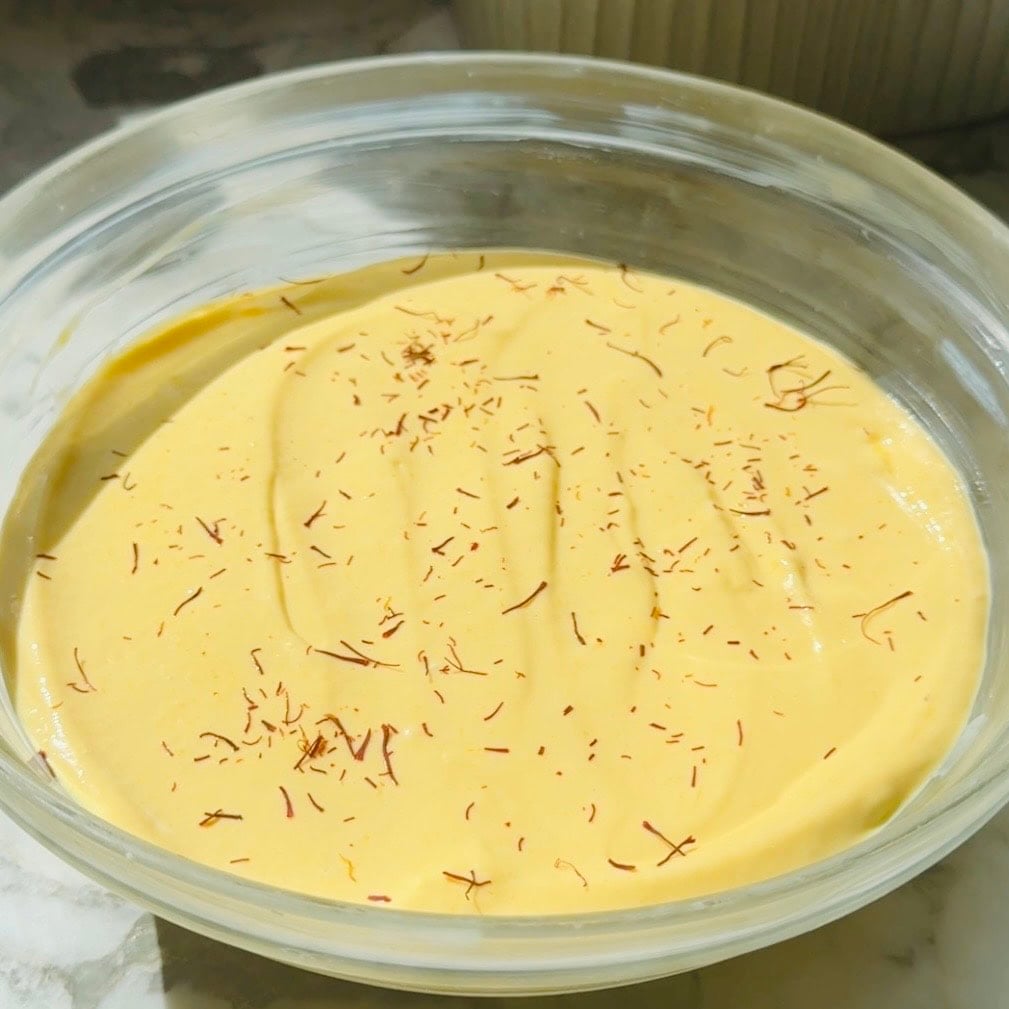 A glass bowl filled with thick, yellow saffron-infused indian mango dessert dessert garnished with saffron strands on top.
