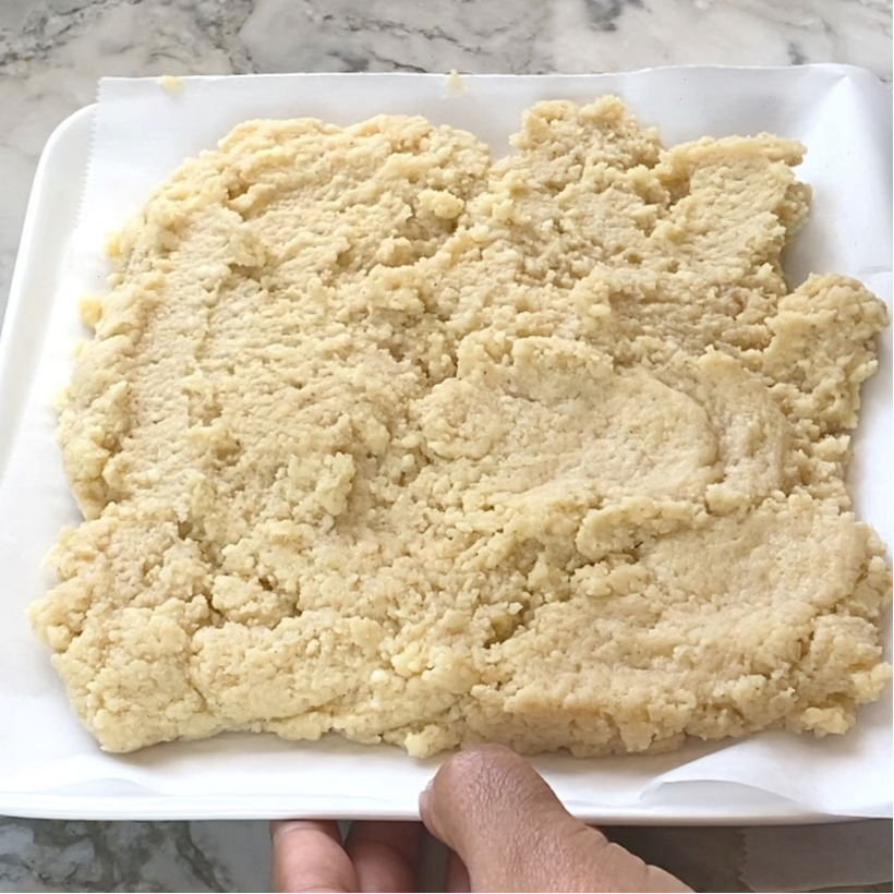 A hand holds a tray with a rough, spread-out layer of dough or batter on parchment paper, set on a marble countertop.