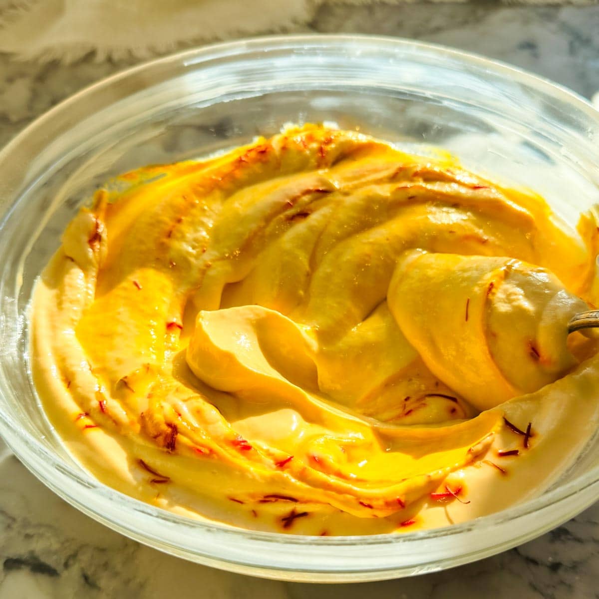 A clear glass bowl filled with creamy, yellow saffron-infused mango shrikhand, swirled with visible saffron strands, on a marble surface.