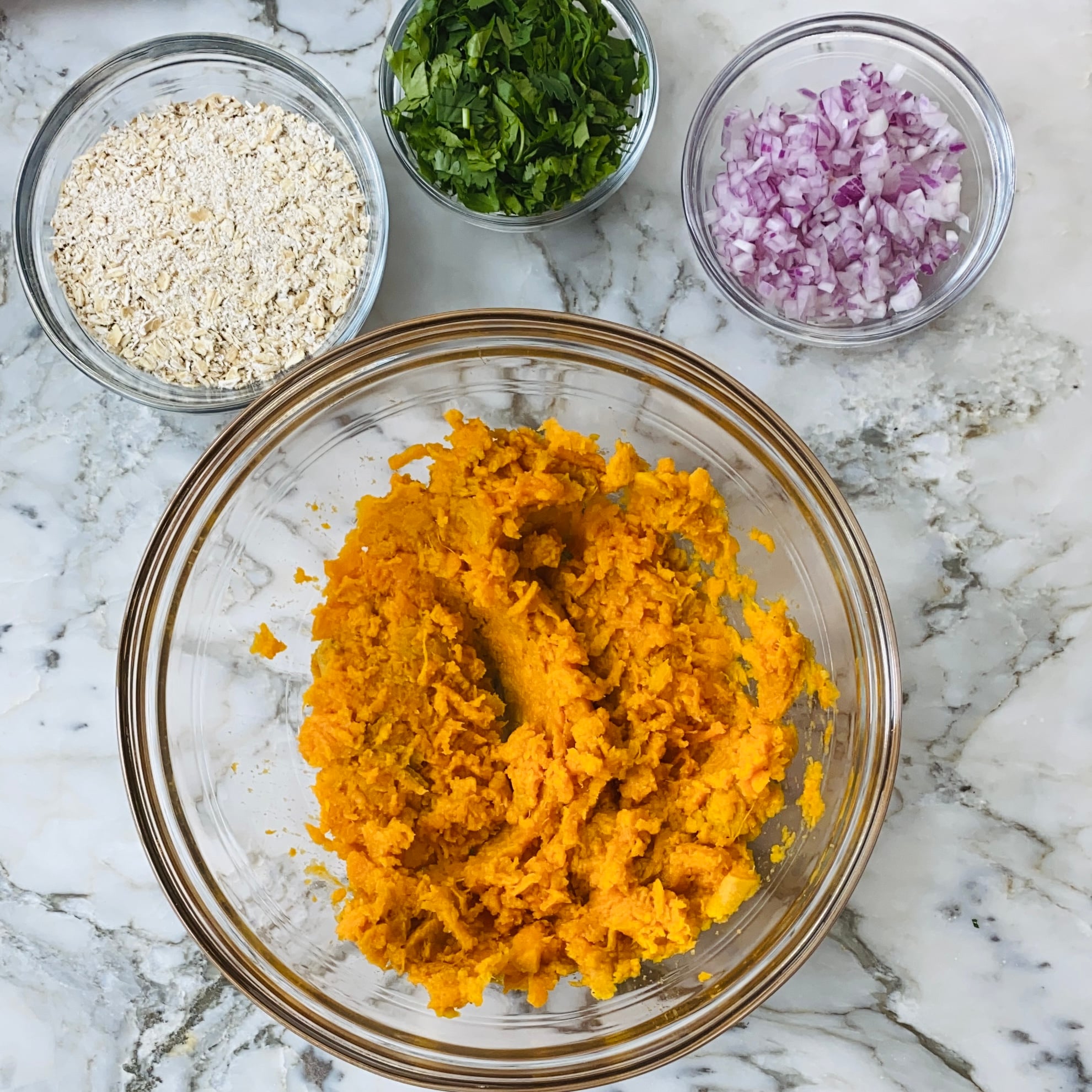 A glass bowl with mashed sweet potato, surrounded by small bowls of chopped red onion, oats, and chopped herbs on a marble countertop.