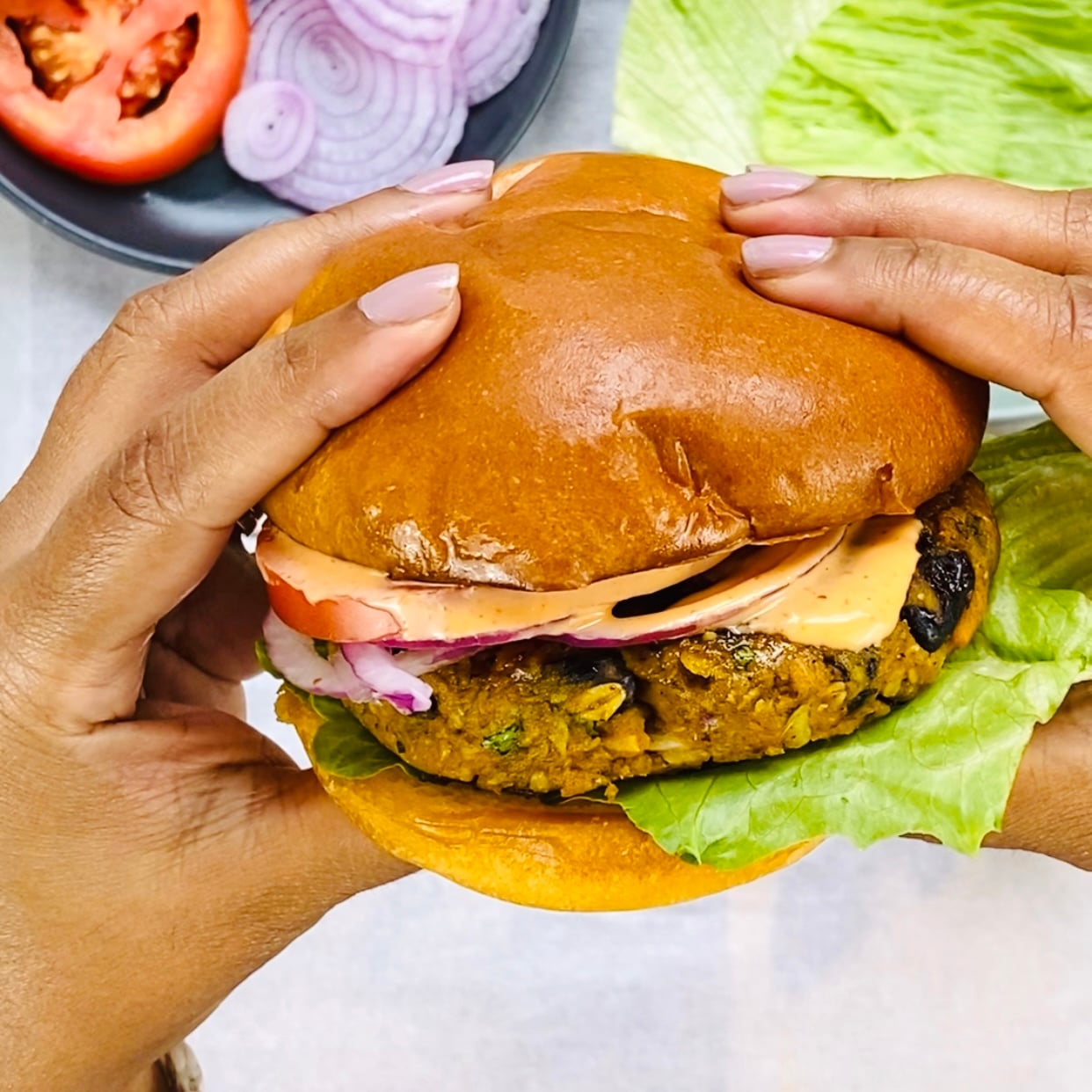 Hands holding a veggie burger with lettuce, tomato, red onion, and sauce in a brioche bun, with sliced vegetables in the background.