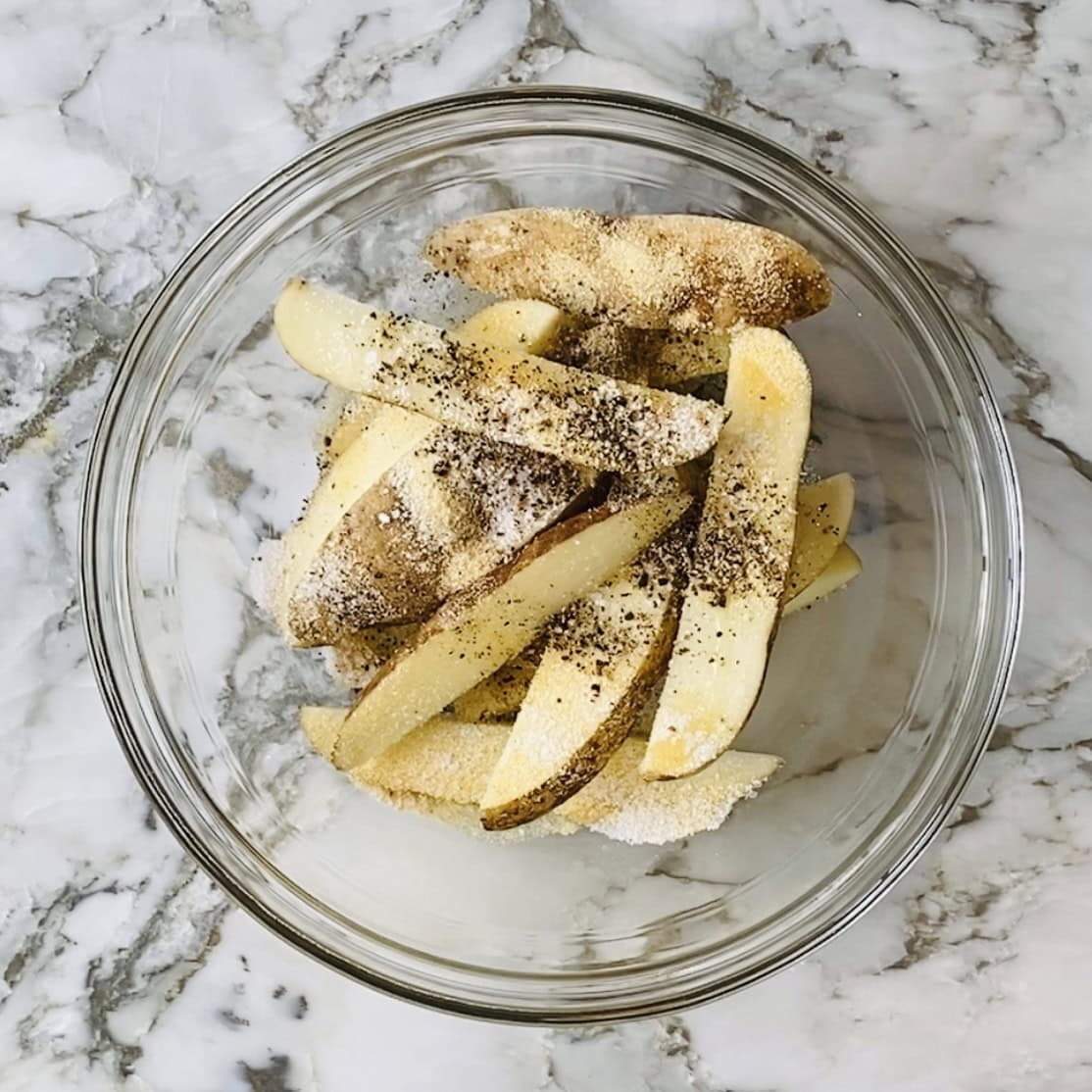 Potato wedges in a glass bowl sprinkled with salt, pepper, and seasoning on a white marble surface.