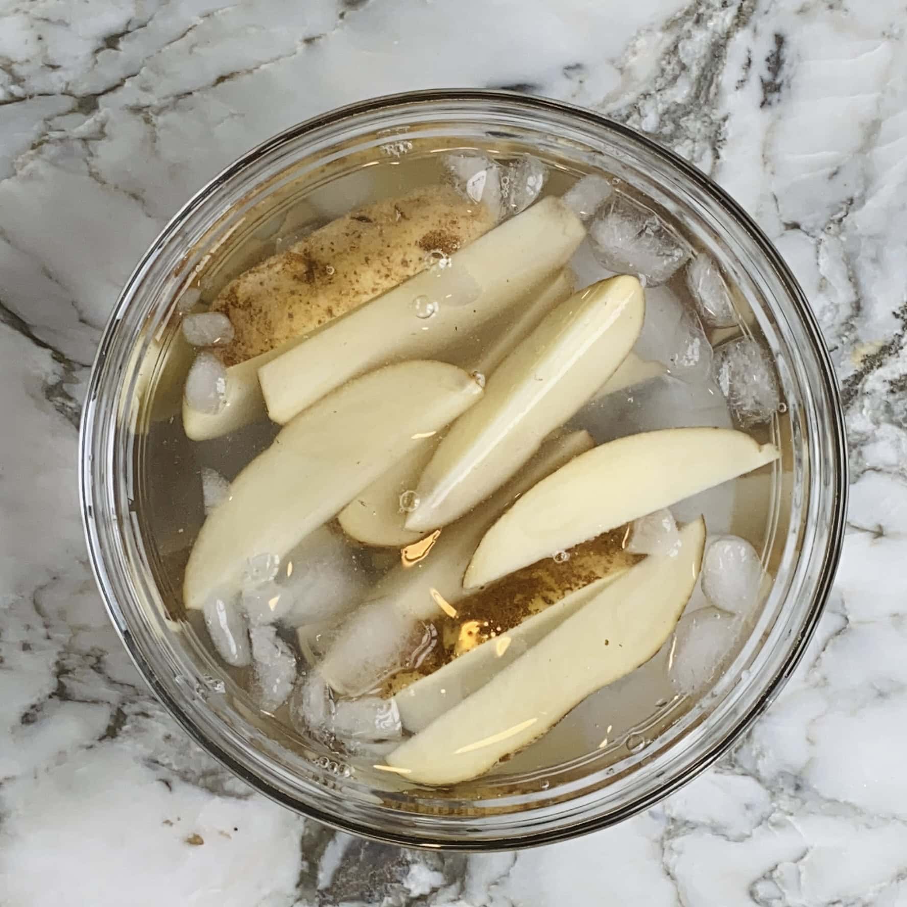 Sliced potatoes soaking in a glass bowl filled with ice water, on a white marble surface.