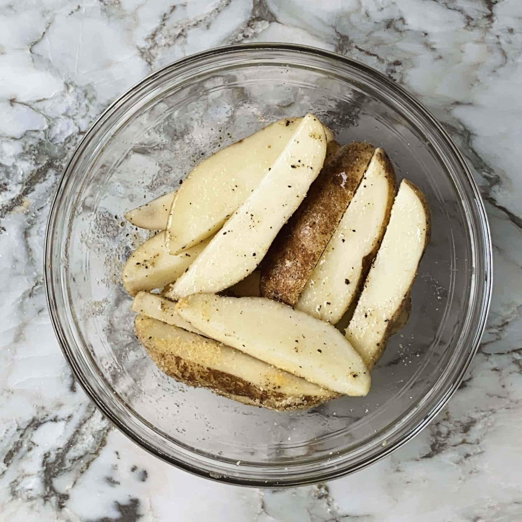 Potato wedges seasoned with salt and pepper in a clear glass bowl on a marble countertop.