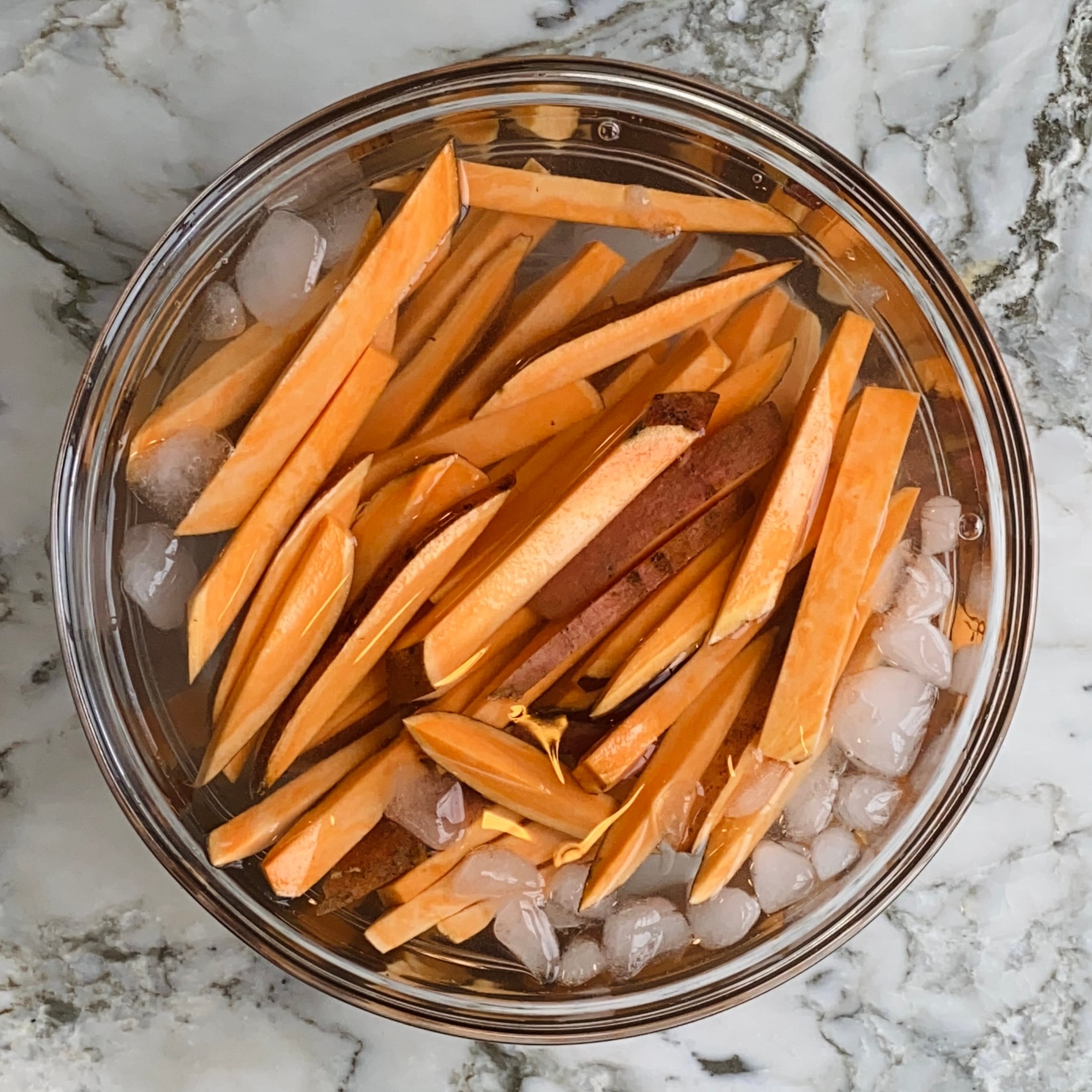 Bowl of raw sweet potato fries soaking in ice water on a marble surface.