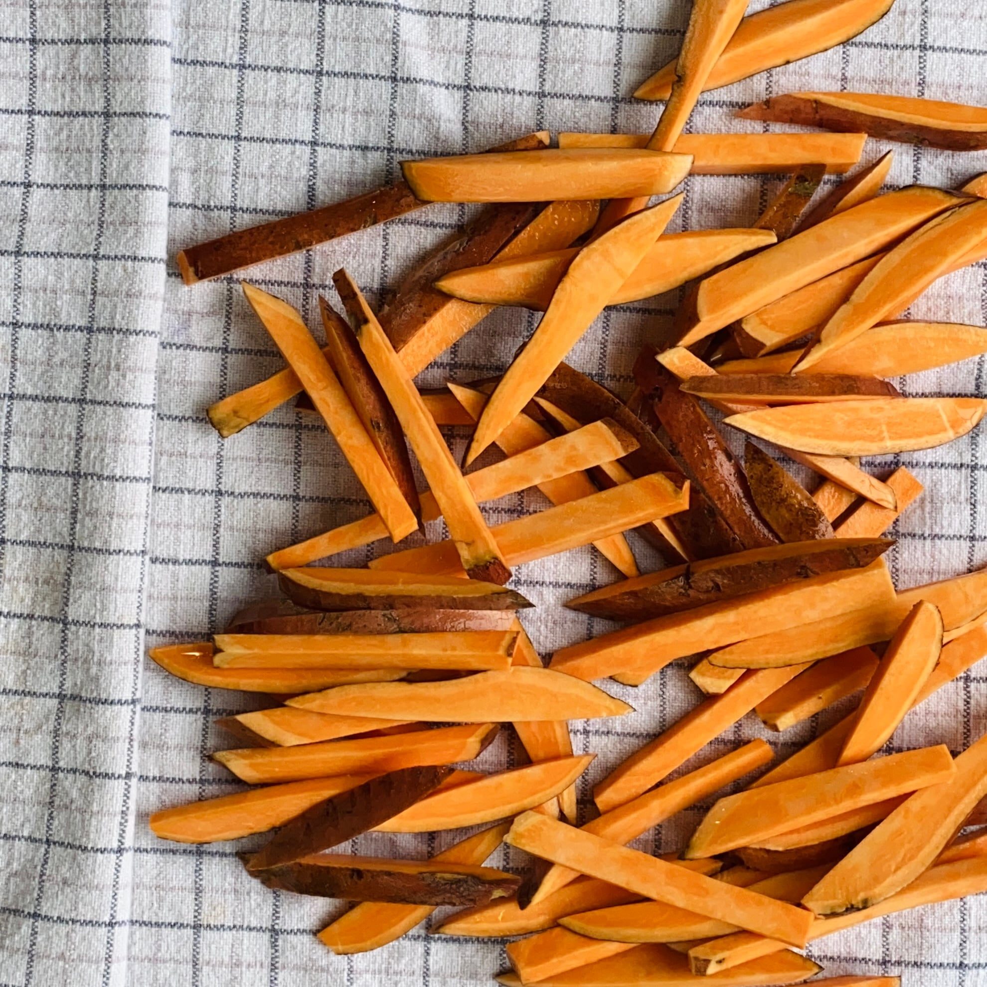 Raw sweet potato fries, skin on, arranged in a pile on a white kitchen towel with a black grid pattern.