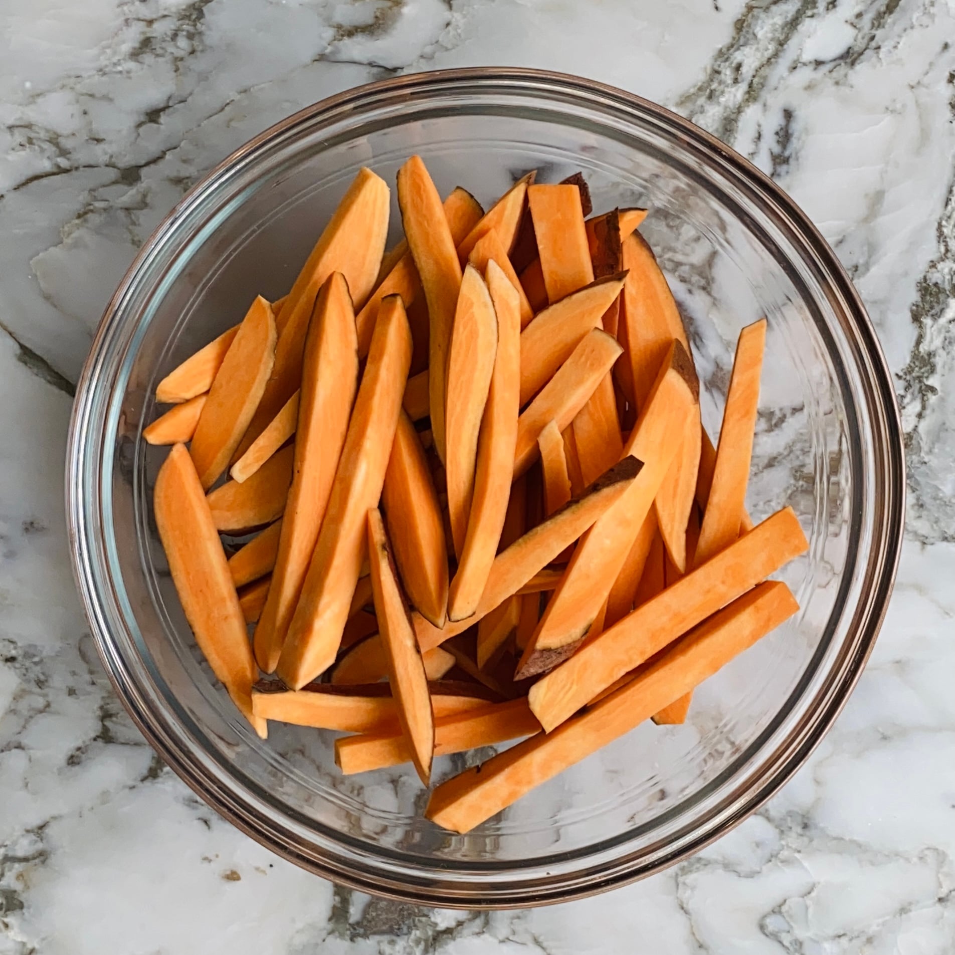 A glass bowl filled with raw sweet potato fries sits on a marble countertop.