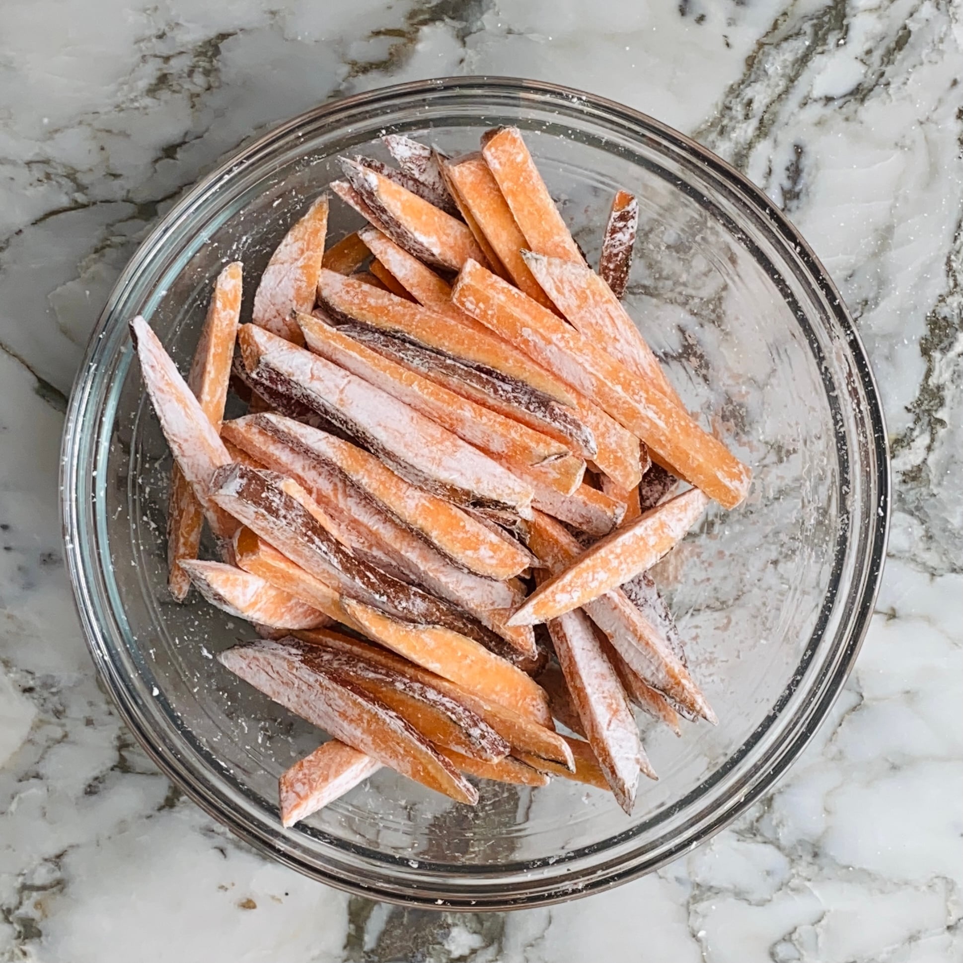 A glass bowl filled with raw sweet potato fries coated in cornstarch sits on a marble countertop.