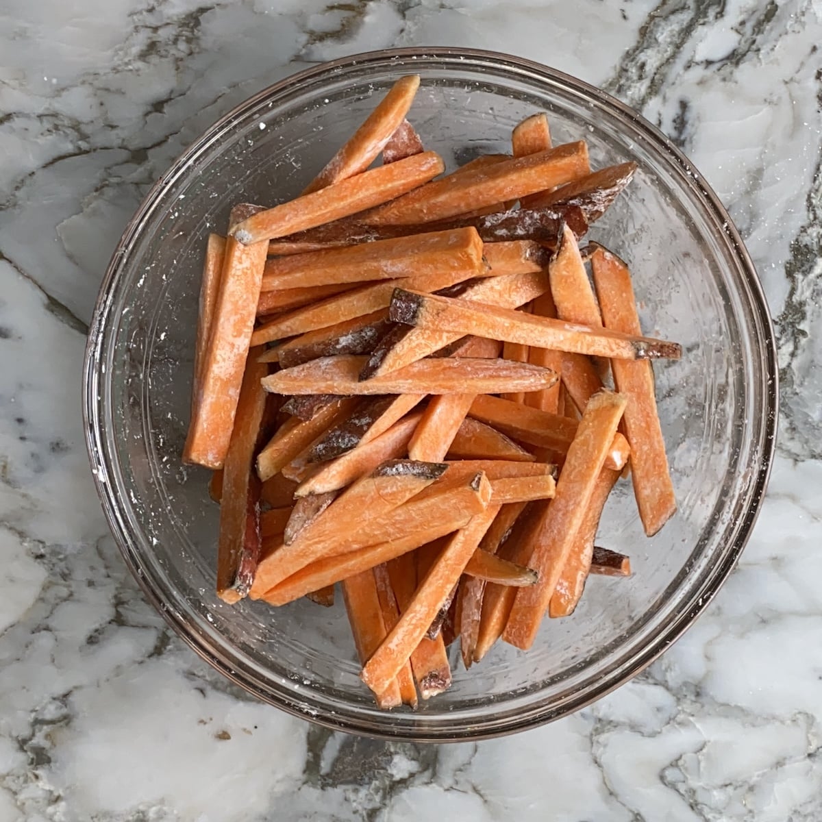 A glass bowl filled with raw sweet potato fries coated in seasoning sits on a marble countertop.