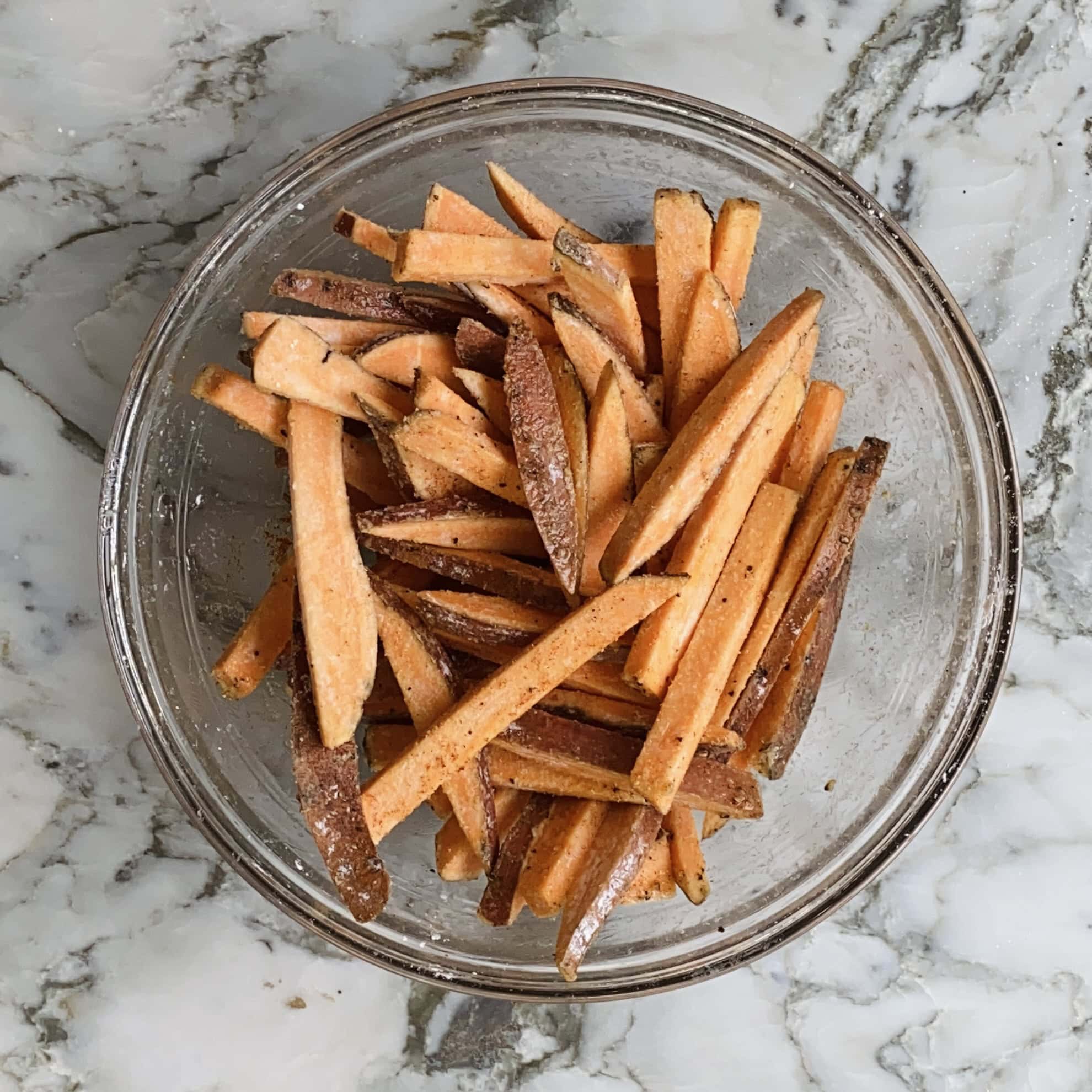 A clear glass bowl containing uncooked, seasoned sweet potato fries sits on a marble surface.
