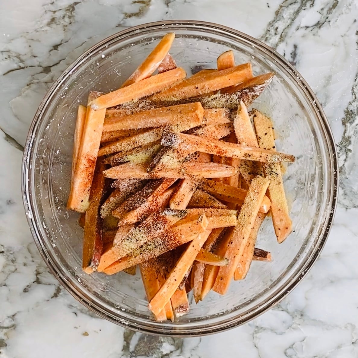 A glass bowl filled with raw sweet potato fries seasoned with various spices sits on a marble countertop.
