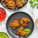 A plate of kanda bhajji (crispy Indian onion fritters) with green chutney, tomato sauce, and cups of chai on a white background.