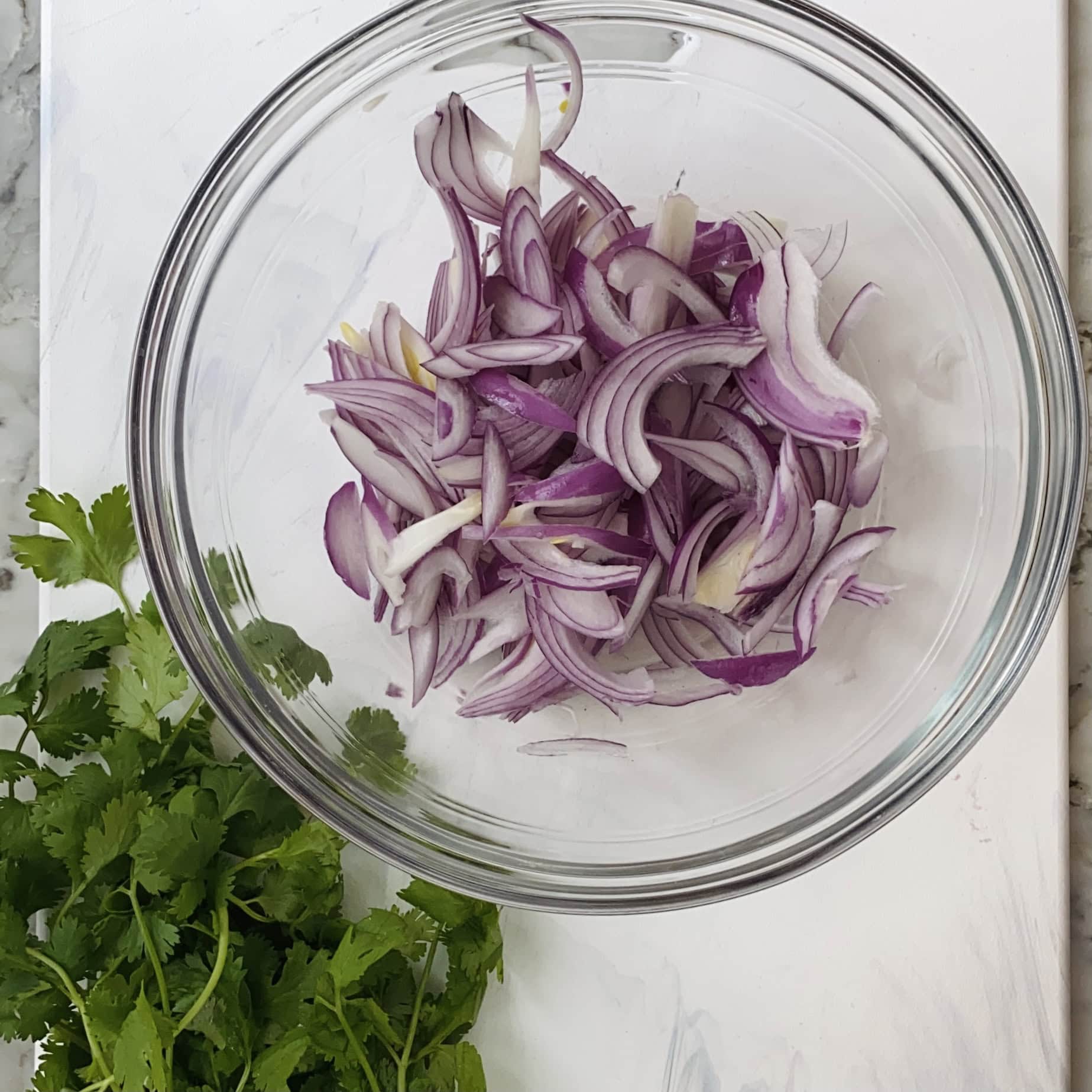 A clear glass bowl containing thinly sliced red onions sits on a white surface next to a bunch of fresh cilantro.