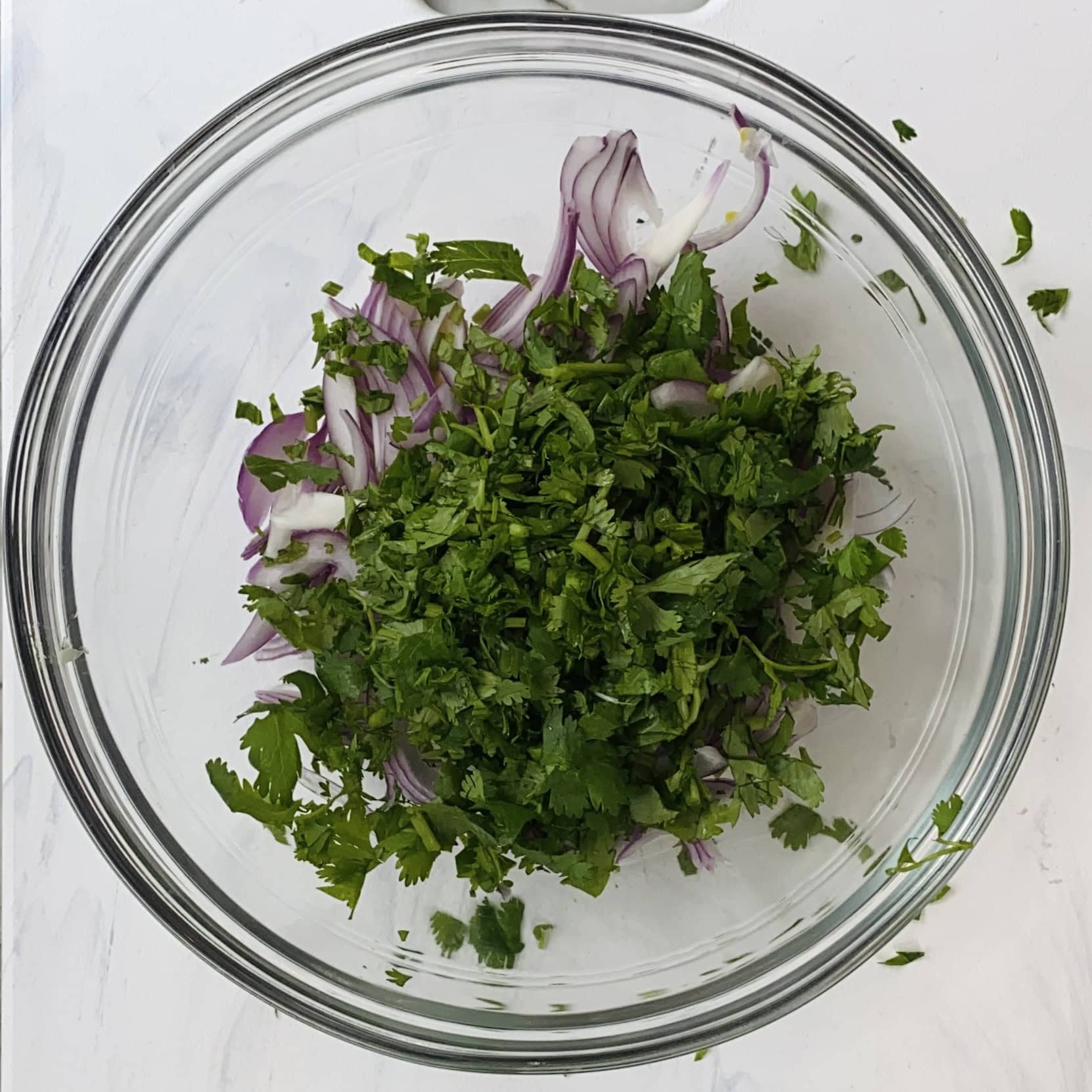 A glass bowl containing chopped cilantro and sliced red onions on a white surface.