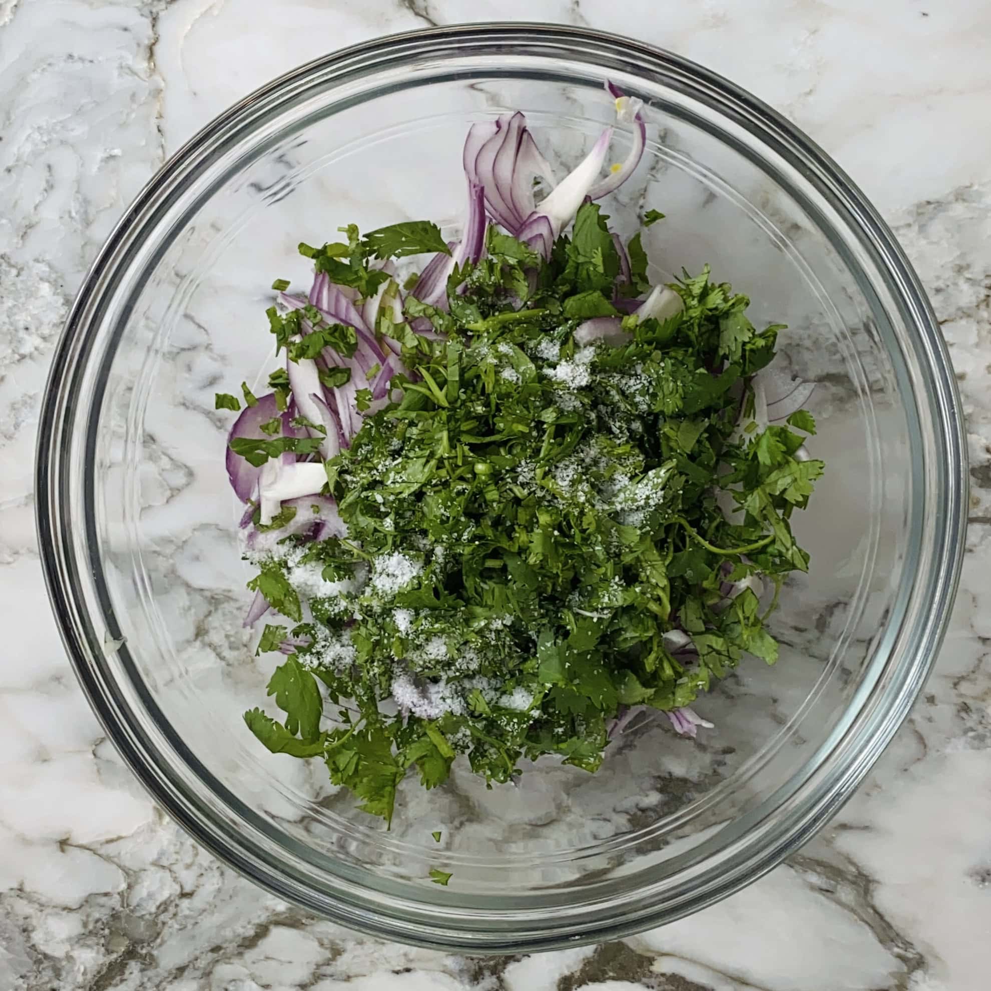 A glass bowl containing sliced red onions, chopped cilantro, and coarse salt on a marble countertop.
