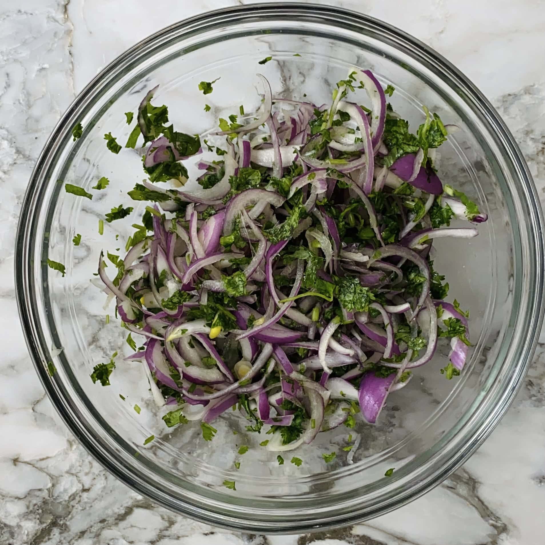 A glass bowl filled with thinly sliced red onions mixed with chopped cilantro on a marble surface.
