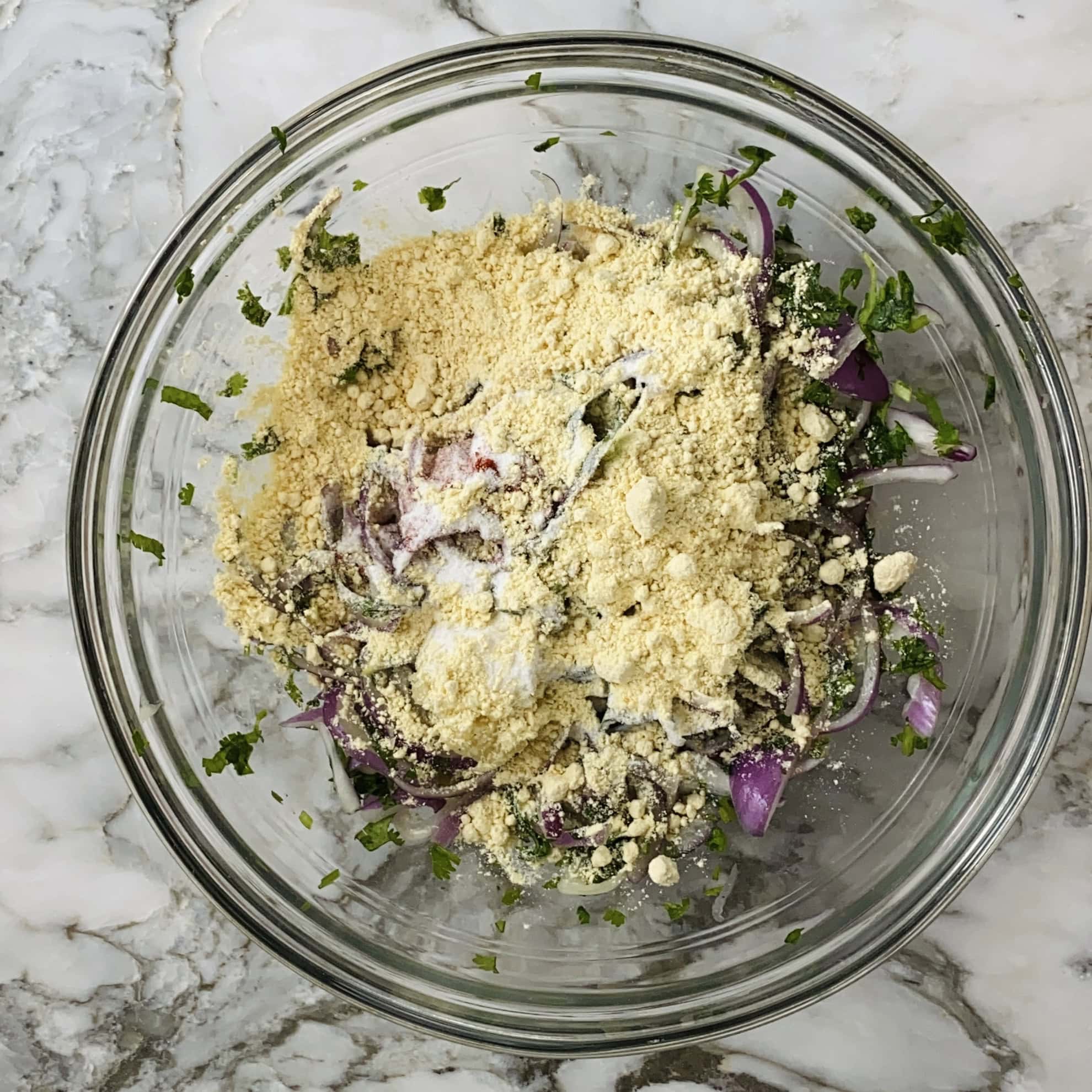 A glass bowl on a marble surface contains sliced red onions, chopped herbs, and chickpea flour being mixed together.