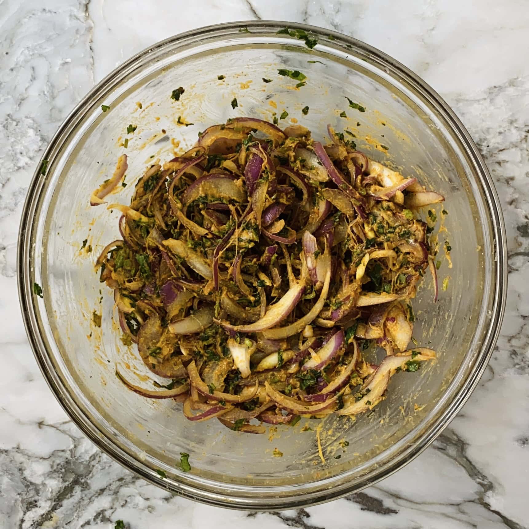 A glass bowl containing sliced red onions mixed with herbs and spices, placed on a marble countertop.