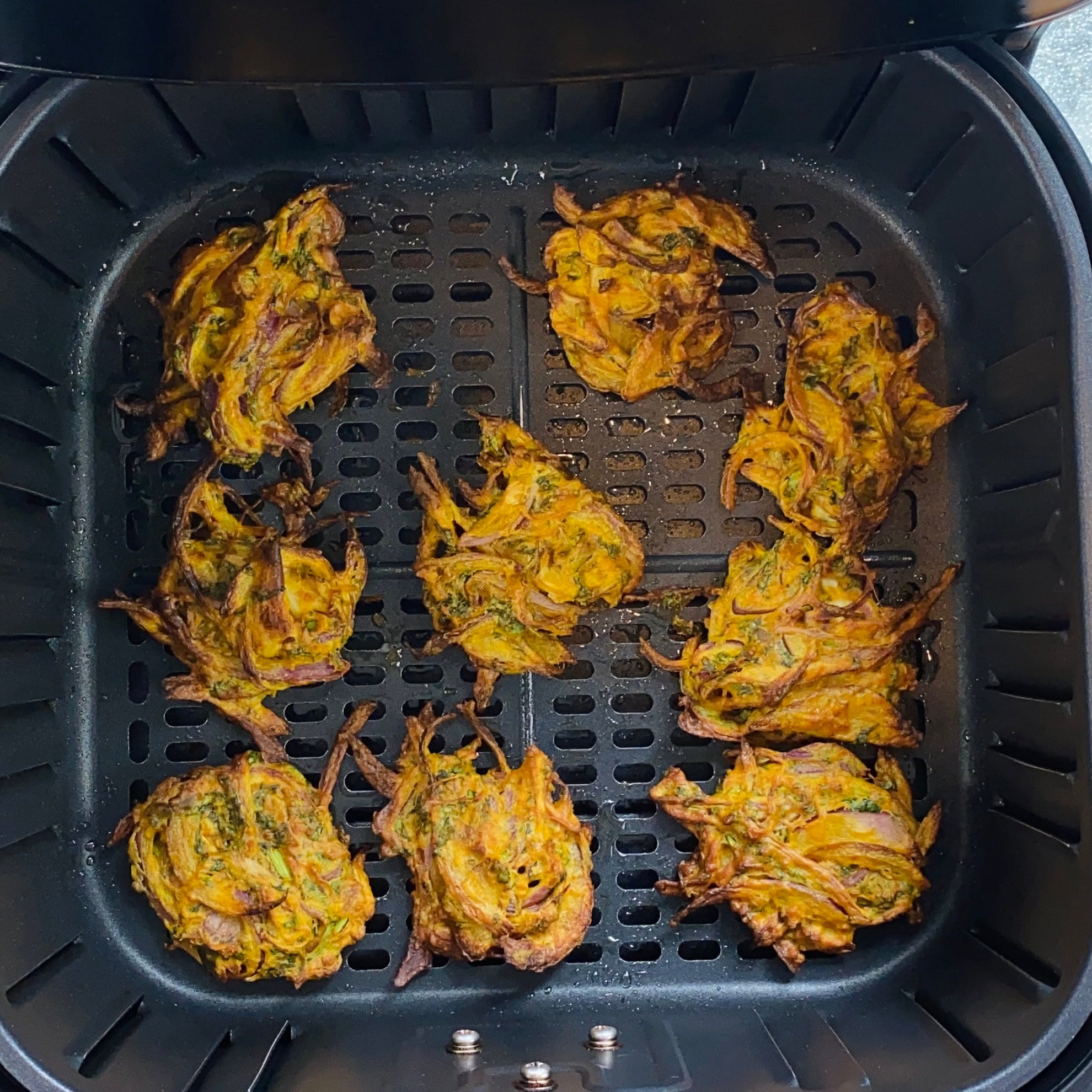 Nine vegetable fritters are arranged in an air fryer basket, partially overlapping, with visible herbs and a golden-brown, crispy texture.