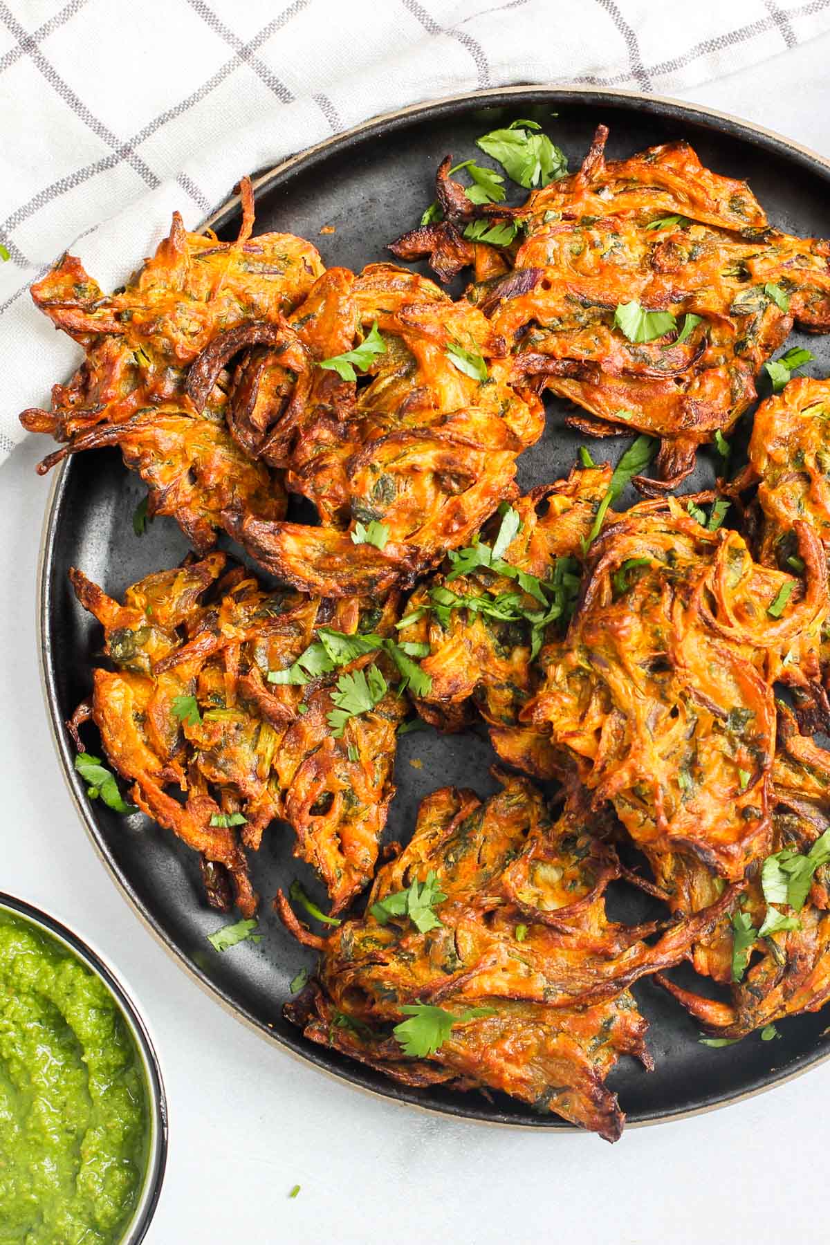 A black plate with several crispy onion bhajis garnished with chopped cilantro, next to a small bowl of green chutney.