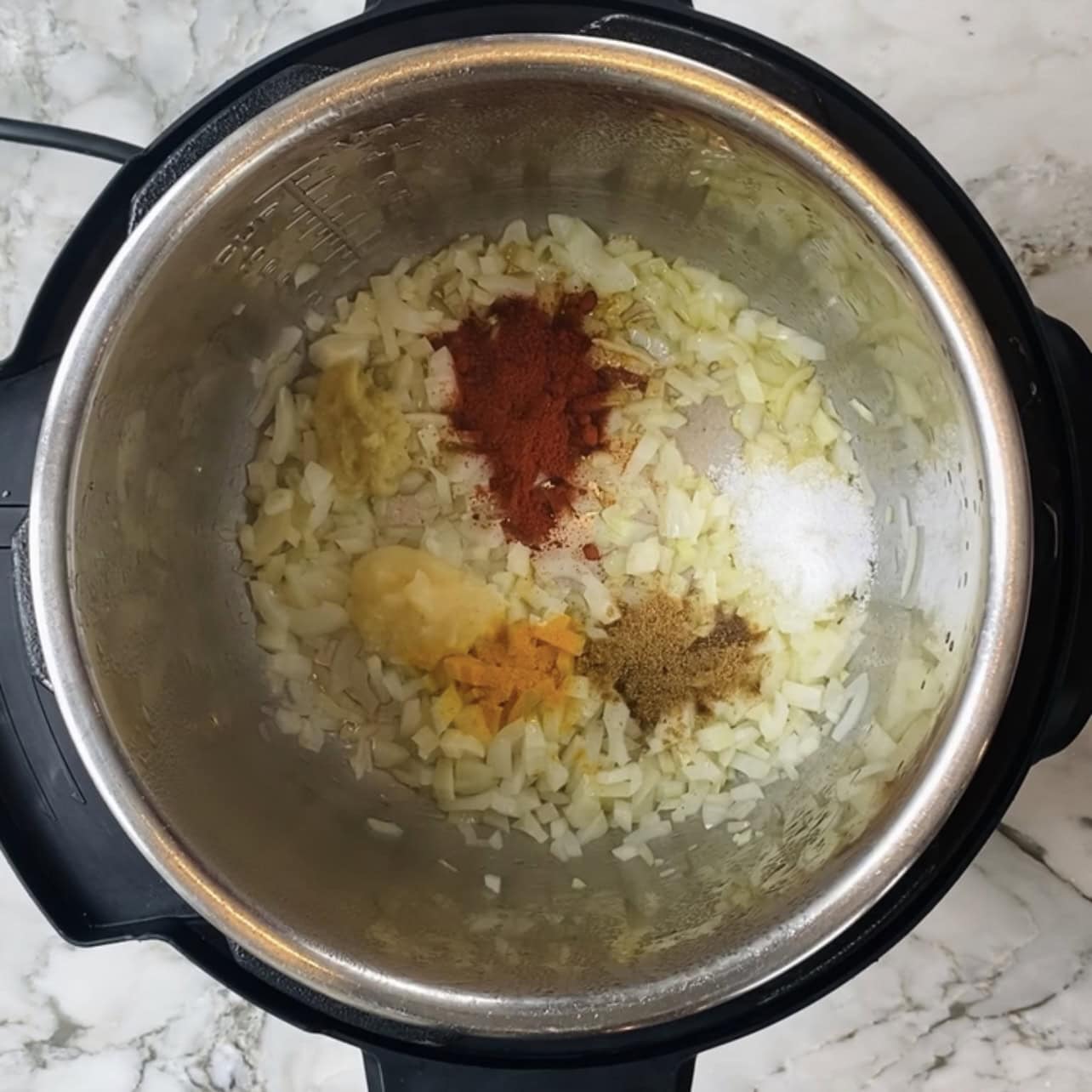 Chopped onions and various ground spices, including turmeric and paprika, in an Instant Pot on a marble countertop.