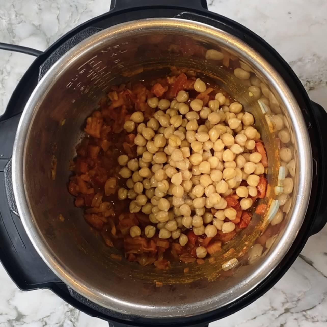 An overhead view of chickpeas and tomato-based ingredients inside an Instant Pot on a marble countertop.