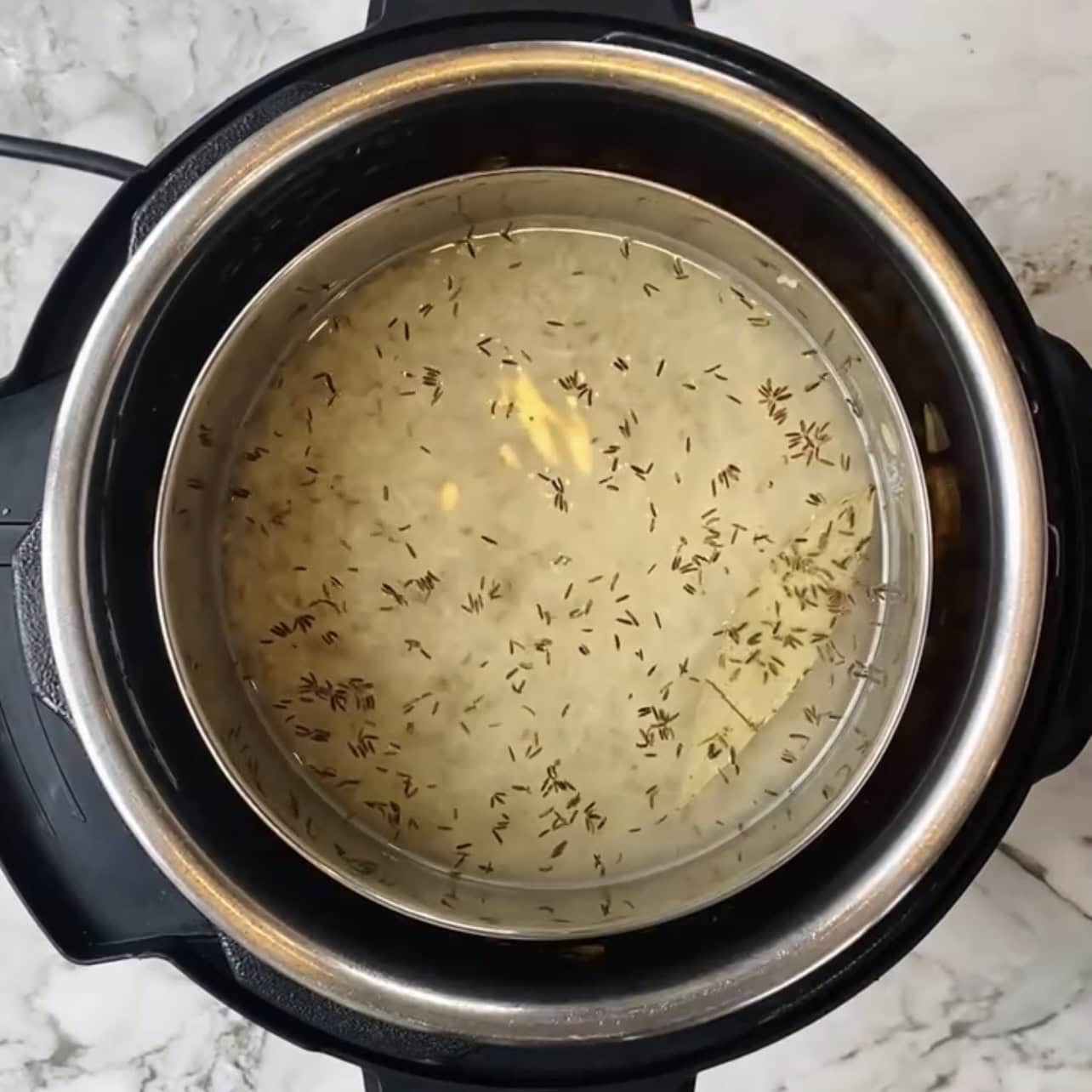 A top view of an Instant Pot containing uncooked rice, water, and cumin seeds on a marble countertop.