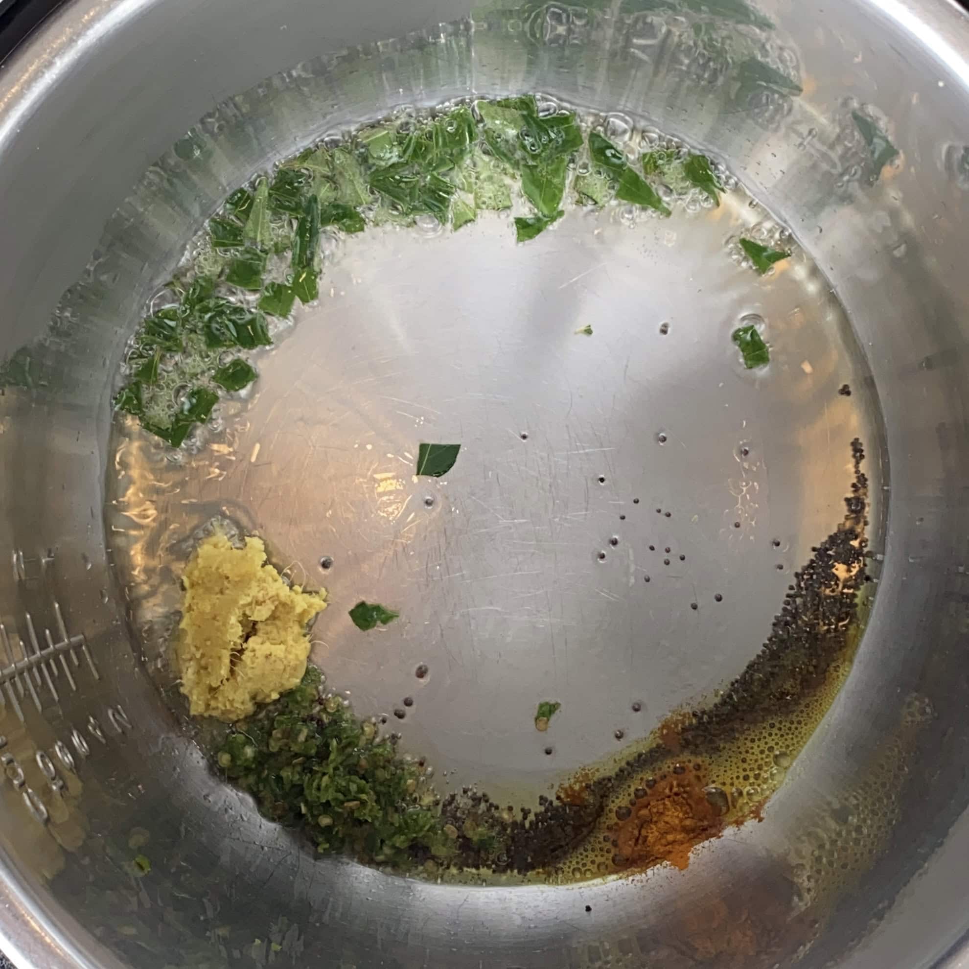 Chopped herbs, ginger paste, mustard seeds, spices, and oil are sizzling in a stainless steel pot.