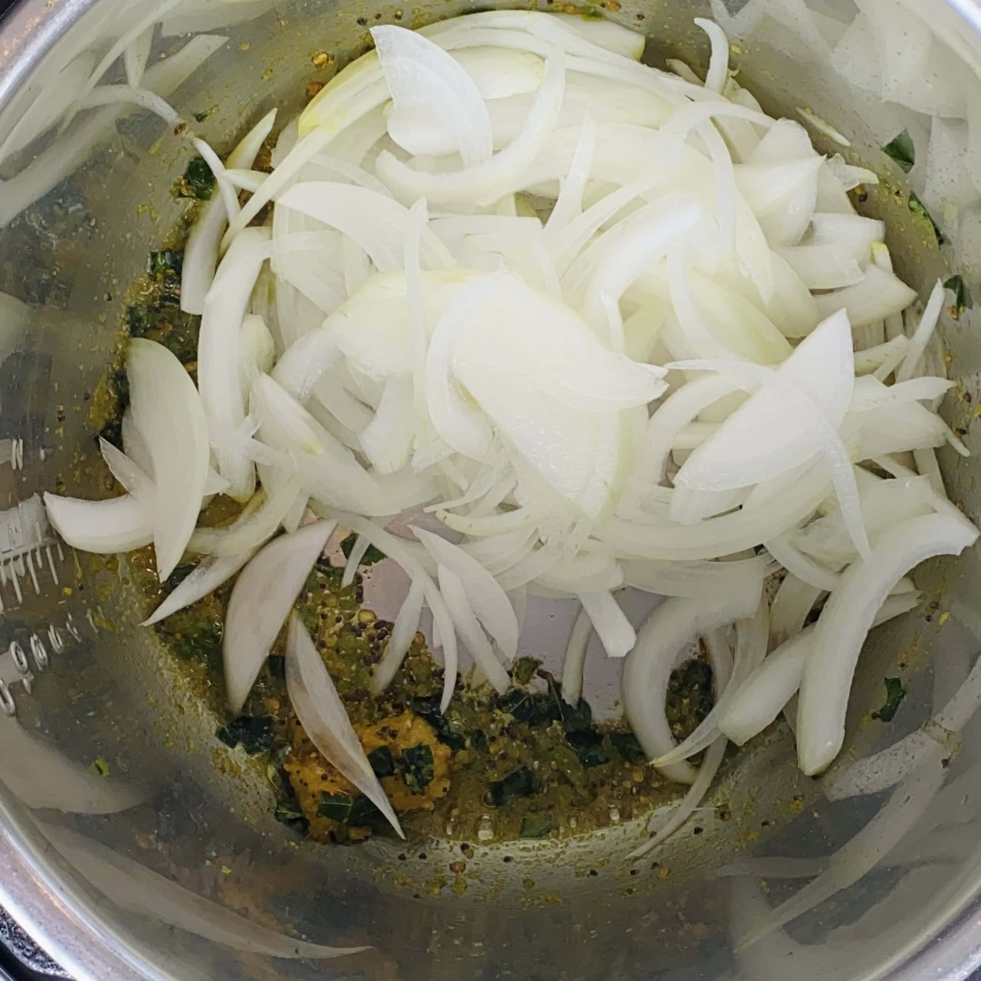 Sliced onions are added to a pot with sautéed spices and chopped herbs at the bottom.