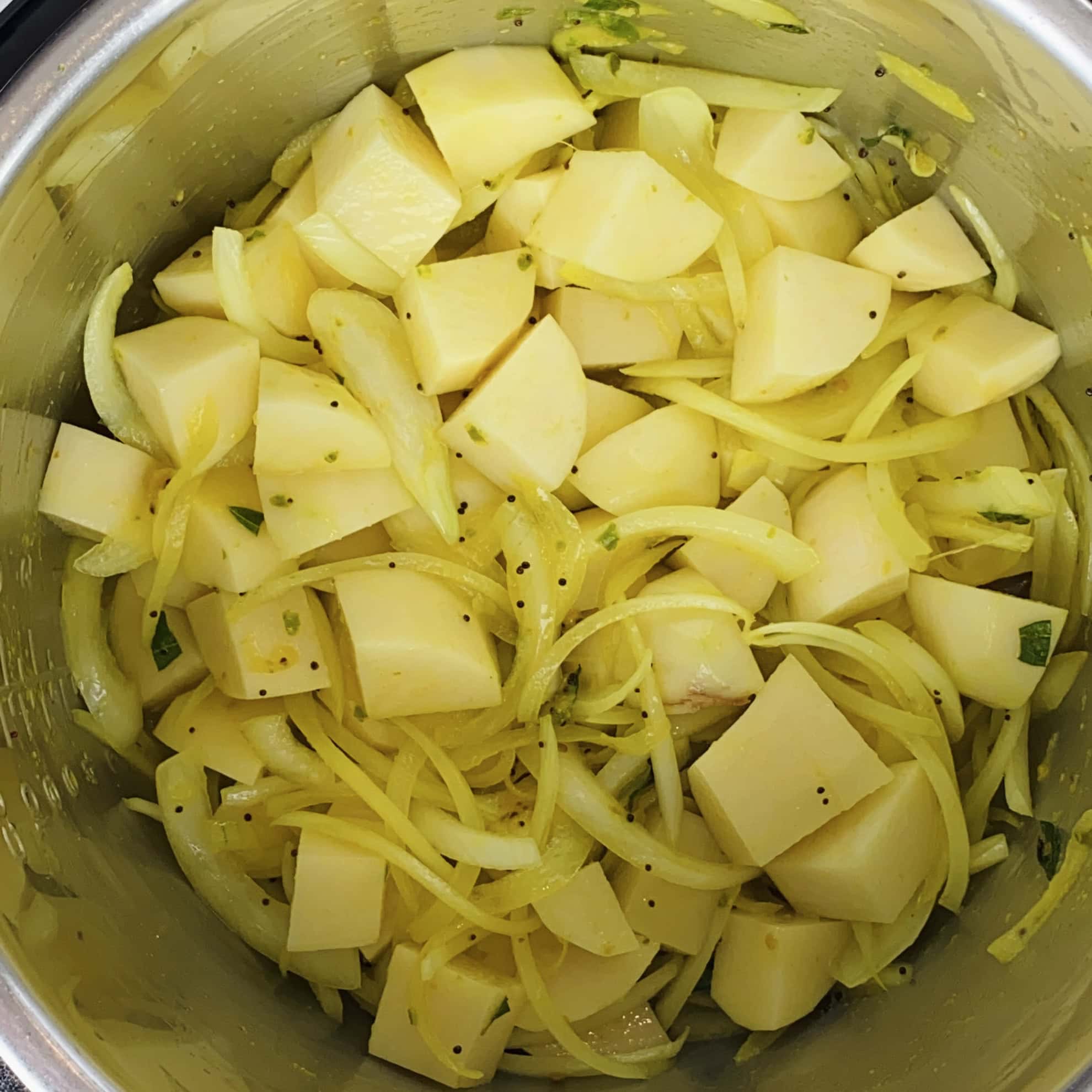 Cubed potatoes and sliced onions mixed with spices in a stainless steel pot, prepared for cooking.