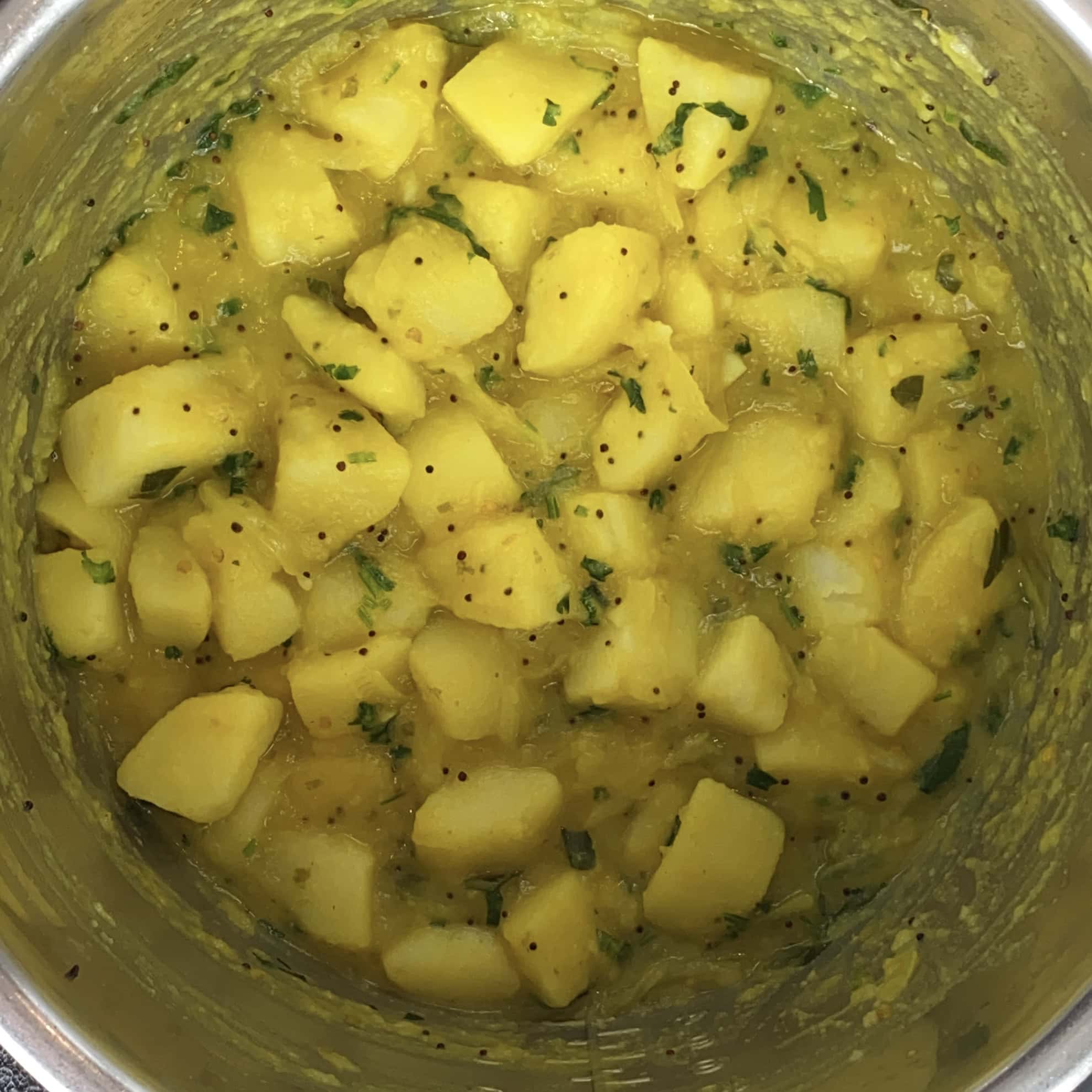 Cubed potatoes cooked in a yellow spiced sauce with visible mustard seeds and chopped herbs in a metal pot.