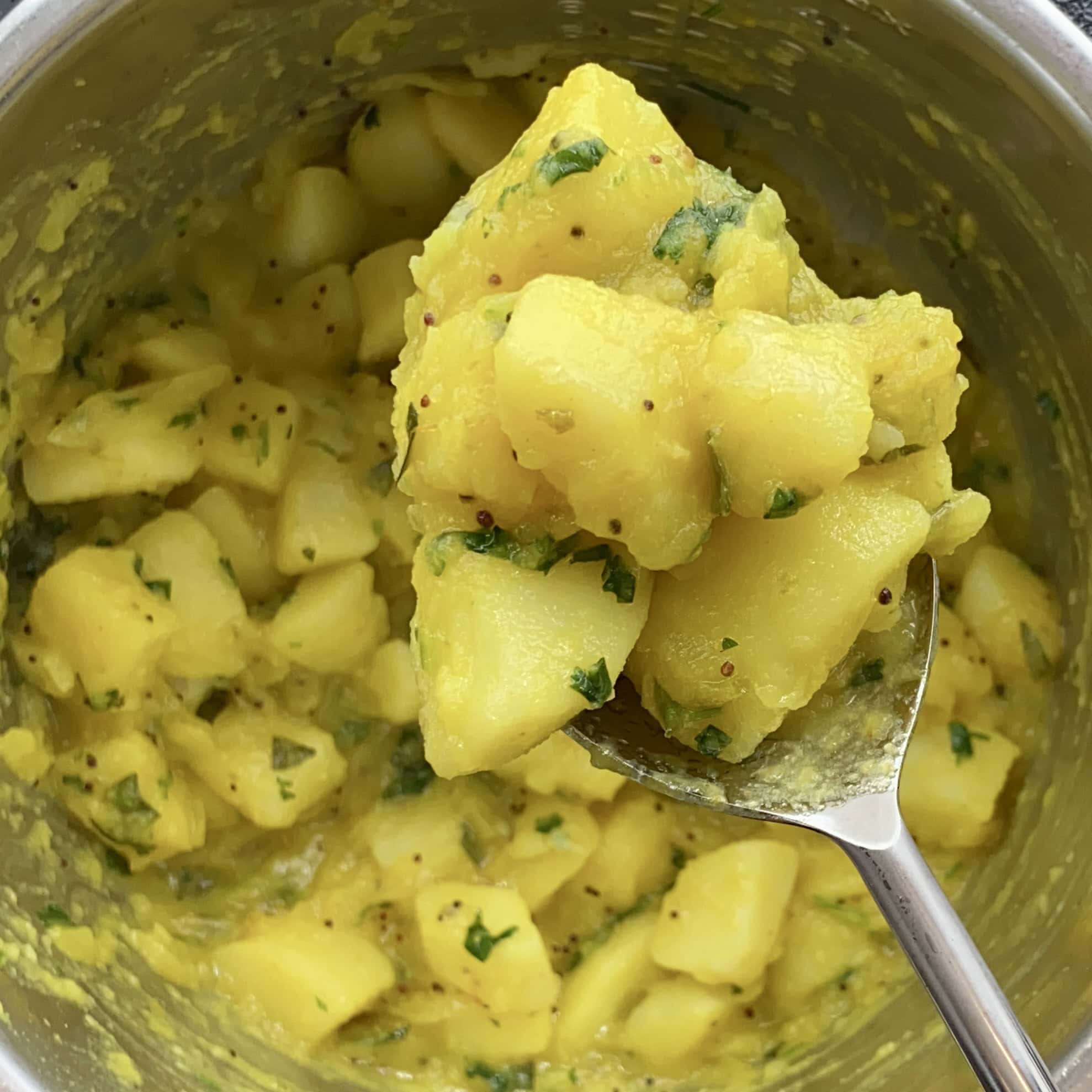 A close-up of a spoonful of yellow, spiced potato cubes garnished with chopped herbs, held above a metal pot containing more of the same dish.