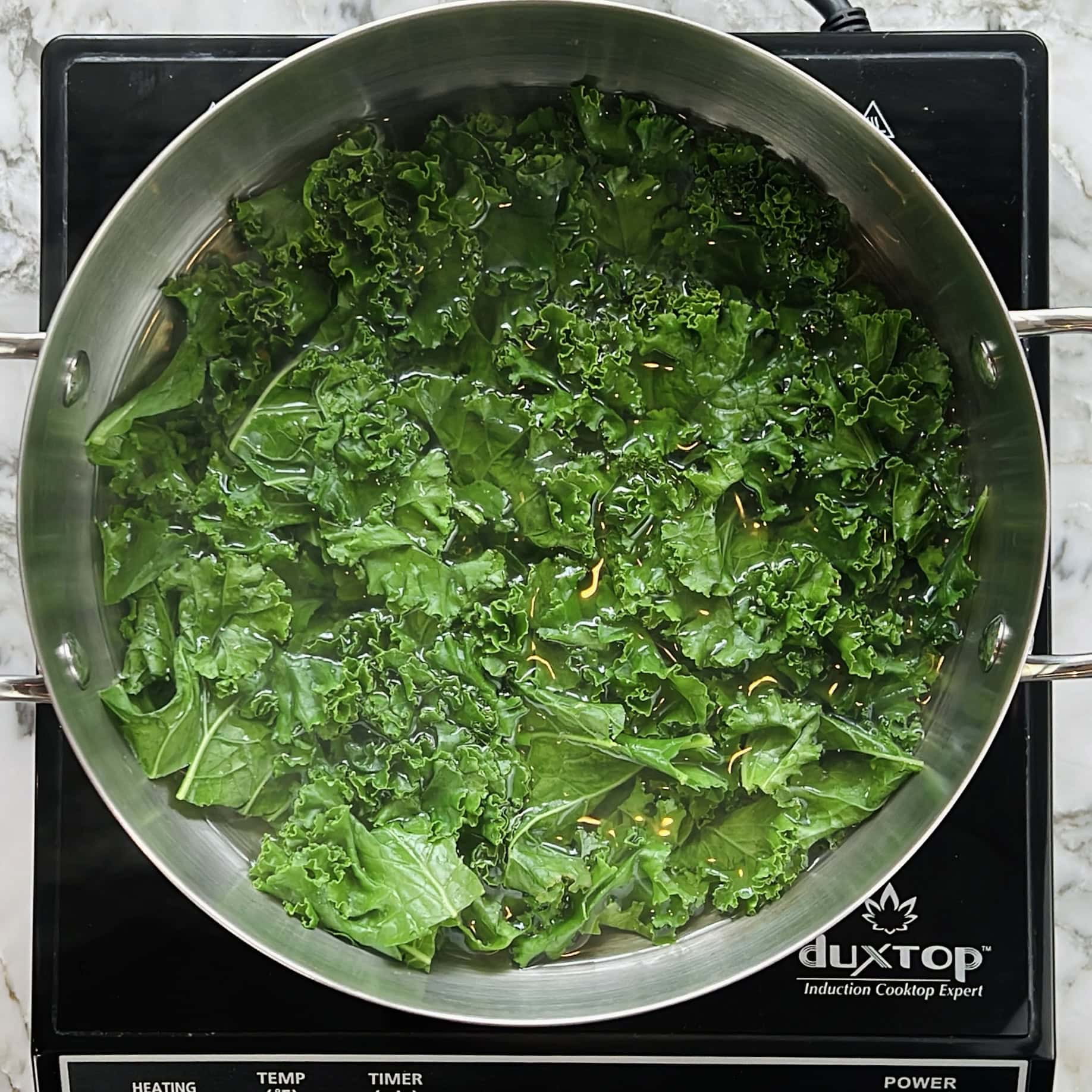 A large pan of fresh kale is being blanched on an induction cooktop.