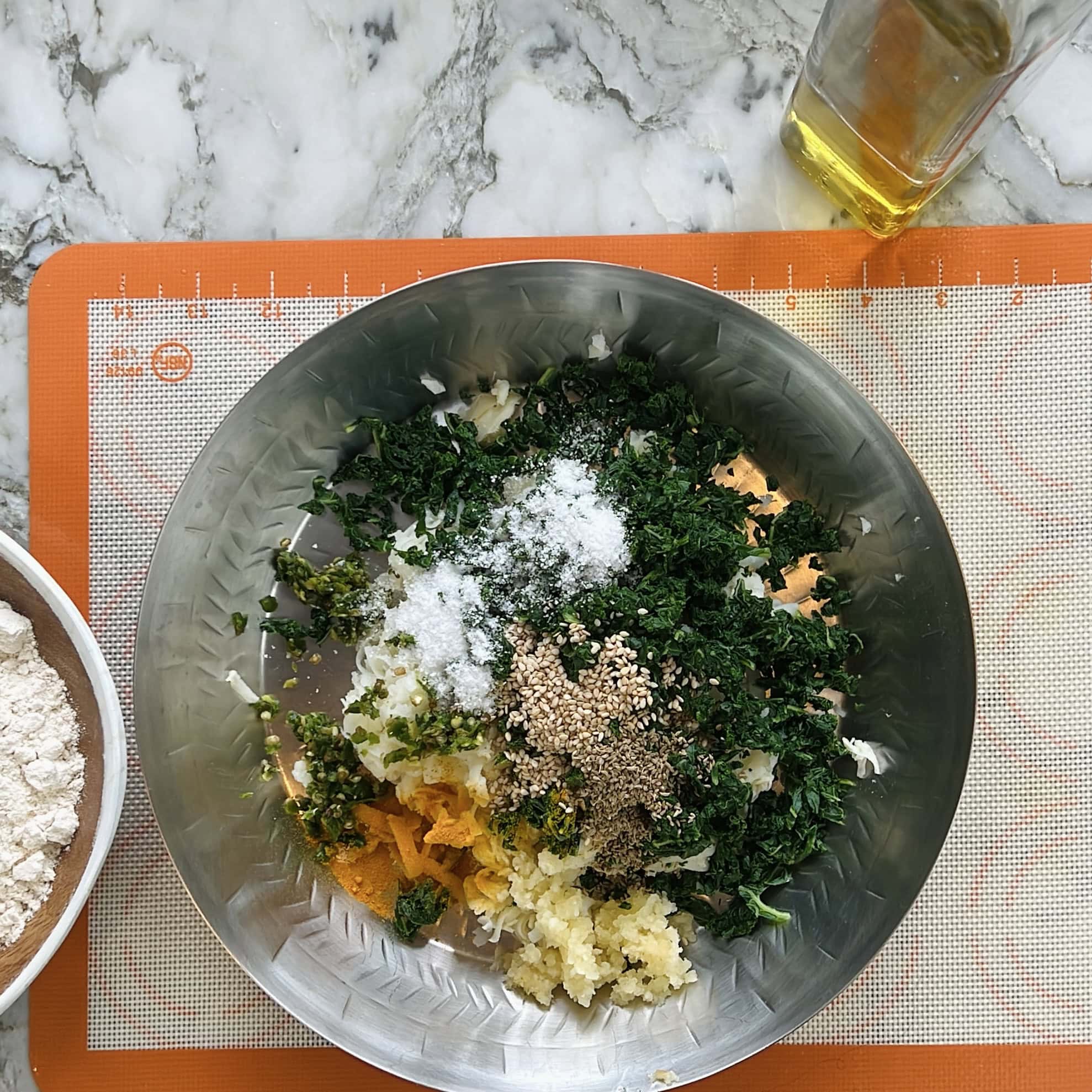 A metal bowl with chopped kale, spices, sesame seeds, and salt sits on a silicone mat next to a bowl of flour and a bottle of oil.